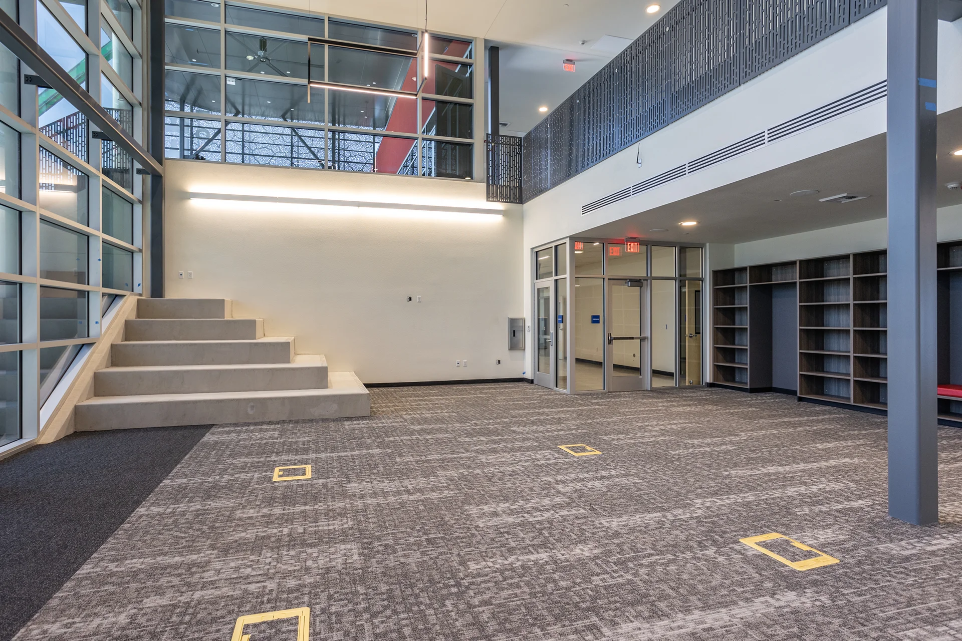 Institutional multi-level common area showing a polished concrete tiered staircase, second-story decorative metal mezzanine railings, glass storefront entryway doors, recessed ceiling lighting, and gray patterned commercial carpeting.