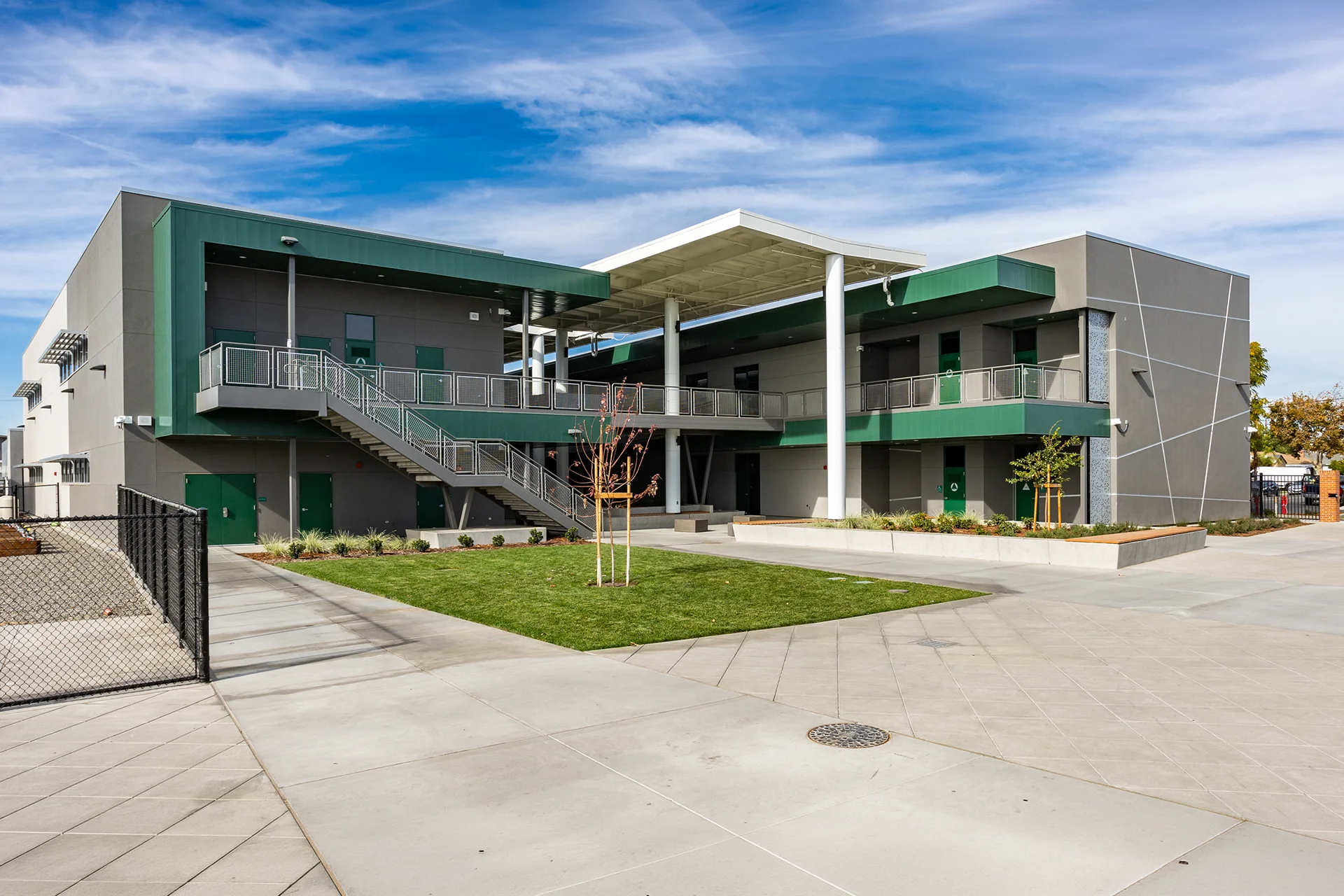 Wide perspective of a modern two-story school courtyard in Manteca, California, showcasing green steel-framed elevated walkways with mesh safety railings, an external concrete staircase, central green lawn area, and a large white architectural canopy supported by structural steel columns.