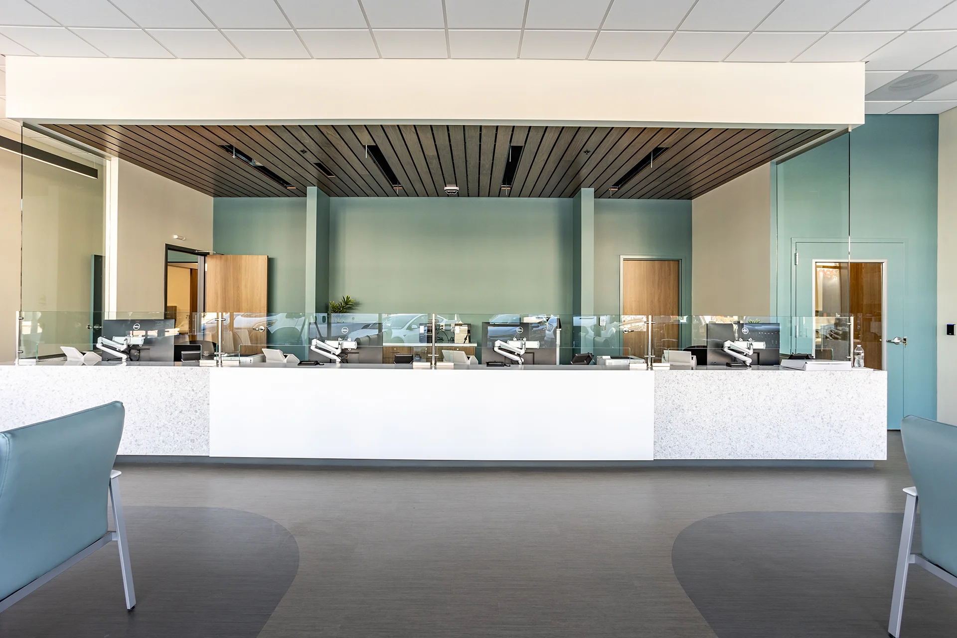 Wide-angle view of a main medical clinic lobby reception desk in San Joaquin County, showcasing custom white terrazzo stone counters, glass privacy sneeze guards, integrated desktop monitors with articulated arms, and a linear dark wood slat ceiling canopy.