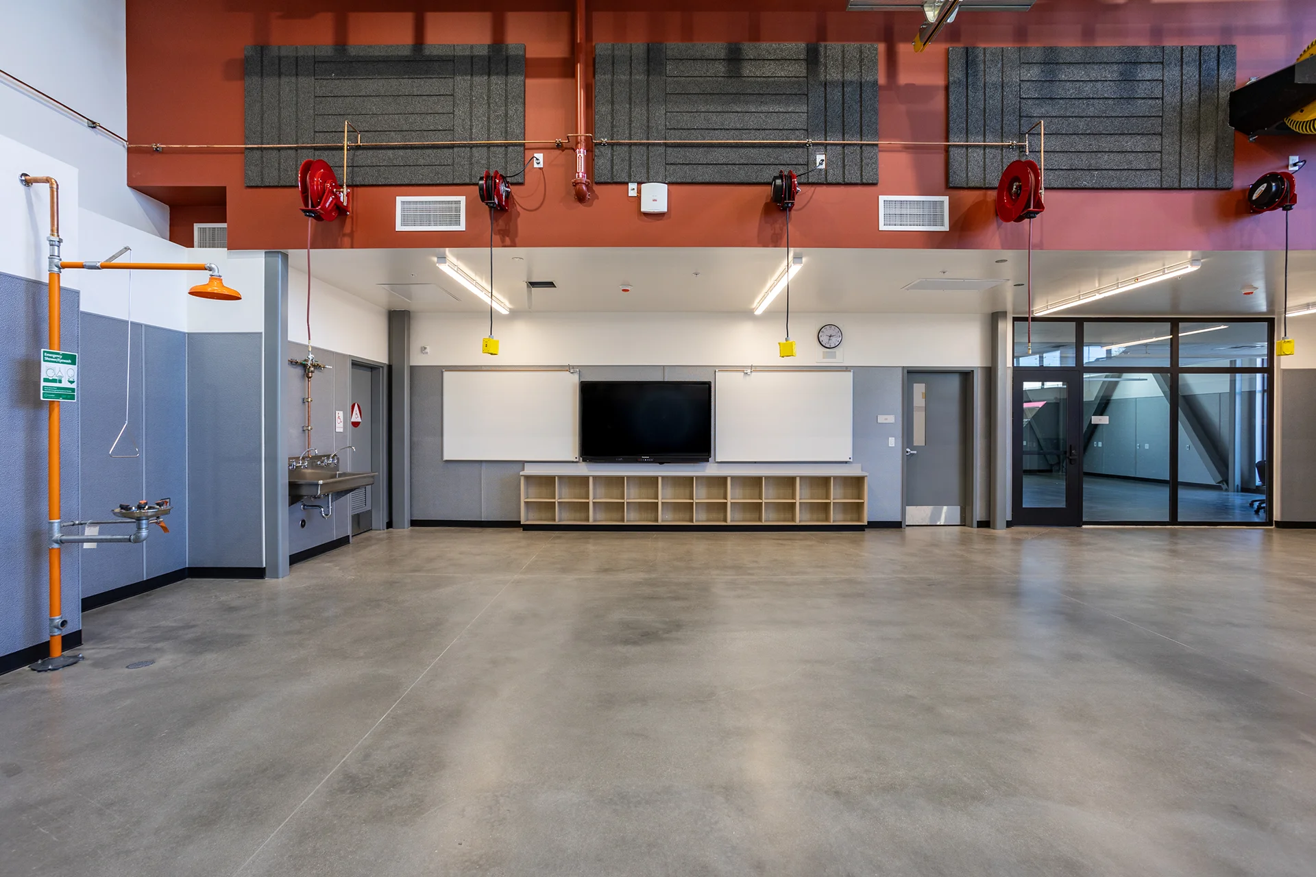 Full-length view of an industrial training lab featuring an integrated eyewash and drench shower safety station, a wall-mounted stainless steel utility sink, overhead drop-down power reels, and acoustic sound-dampening panels fixed to a high red upper bulkhead.