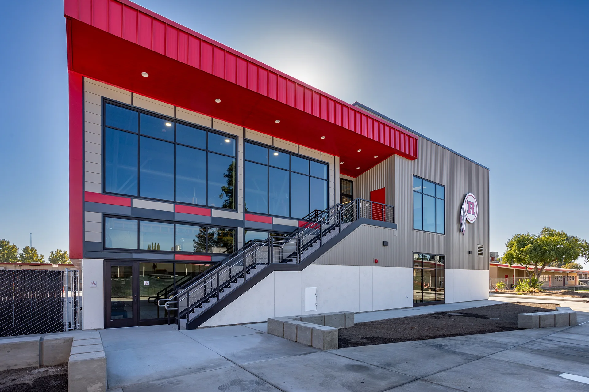 Exterior architectural elevation showcasing large floor-to-ceiling glass curtain walls, a structural steel staircase leading to an upper-level egress platform, a vibrant red modern roof fascia, and architectural concrete block seating.