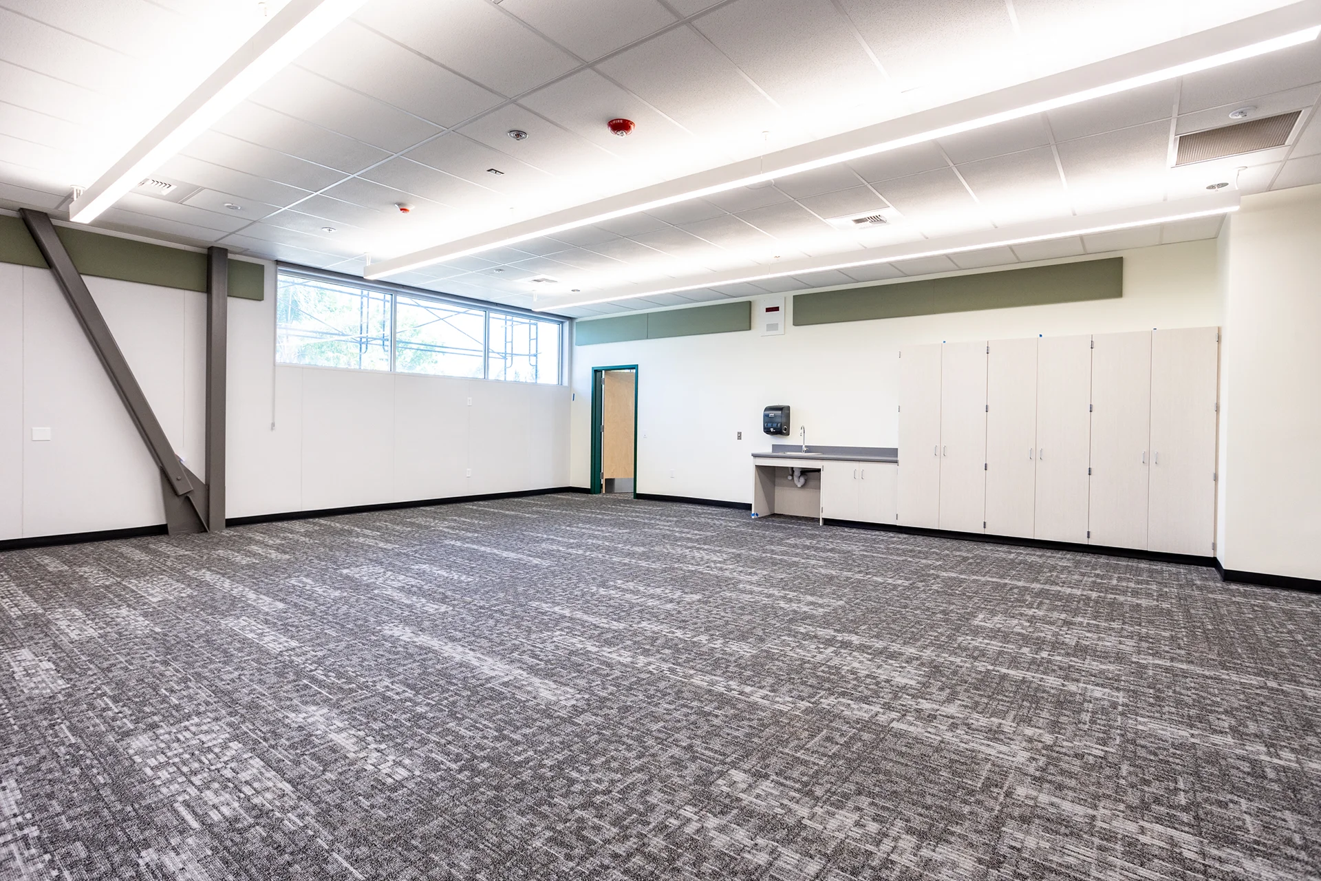 Spacious interior view of an empty classroom space in San Joaquin County, featuring pattern-matched grey commercial carpet tiles, acoustic ceiling tiles with flush-mounted continuous linear LED fixtures, modular wall storage lockers, and a dark grey perimeter wainscoting accent.
