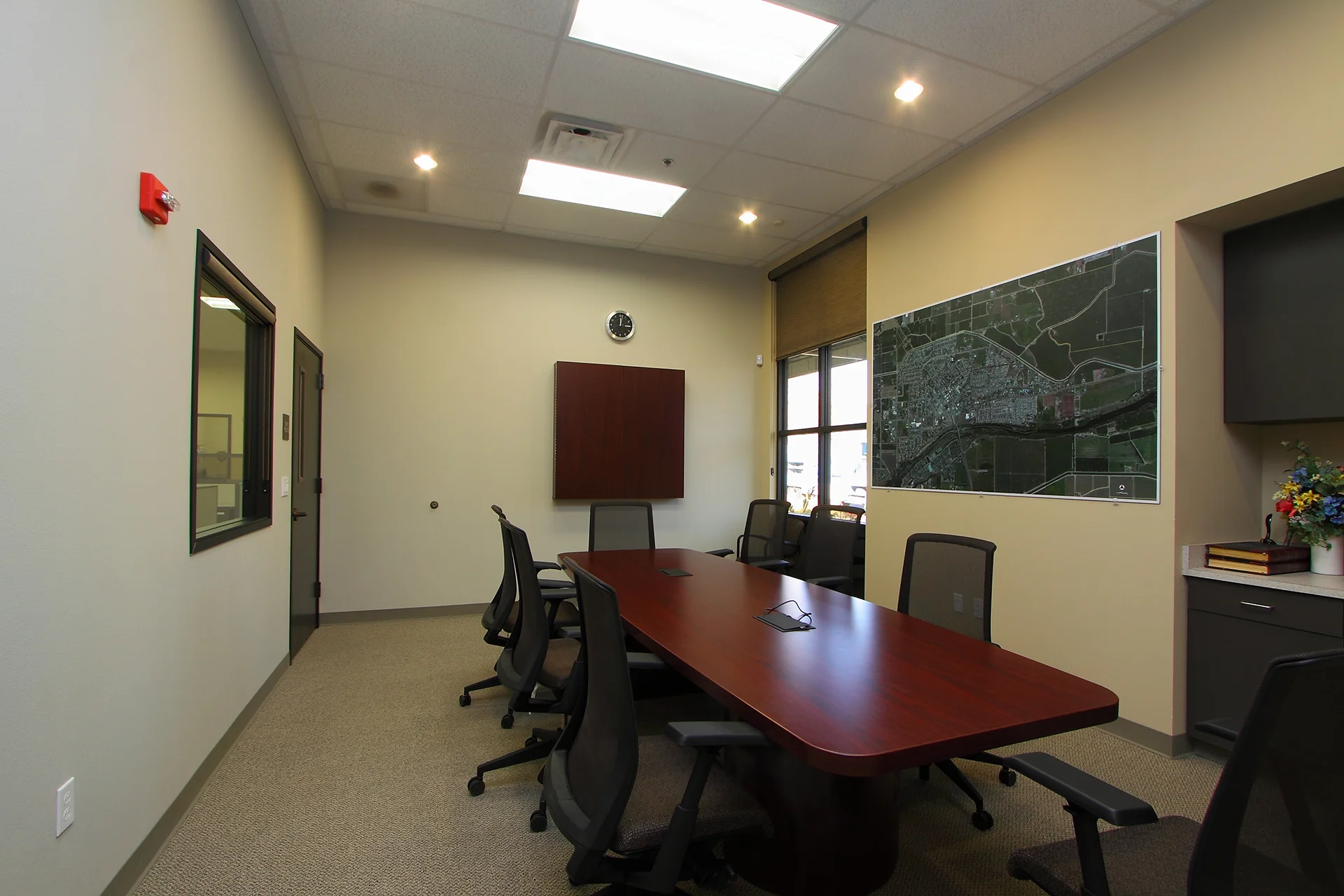 Professional administrative conference room featuring a long polished cherry wood table surrounded by mesh office chairs, an overhead geographic aerial wall map of Waterford, accent lighting, and a service counter with built-in cabinetry.