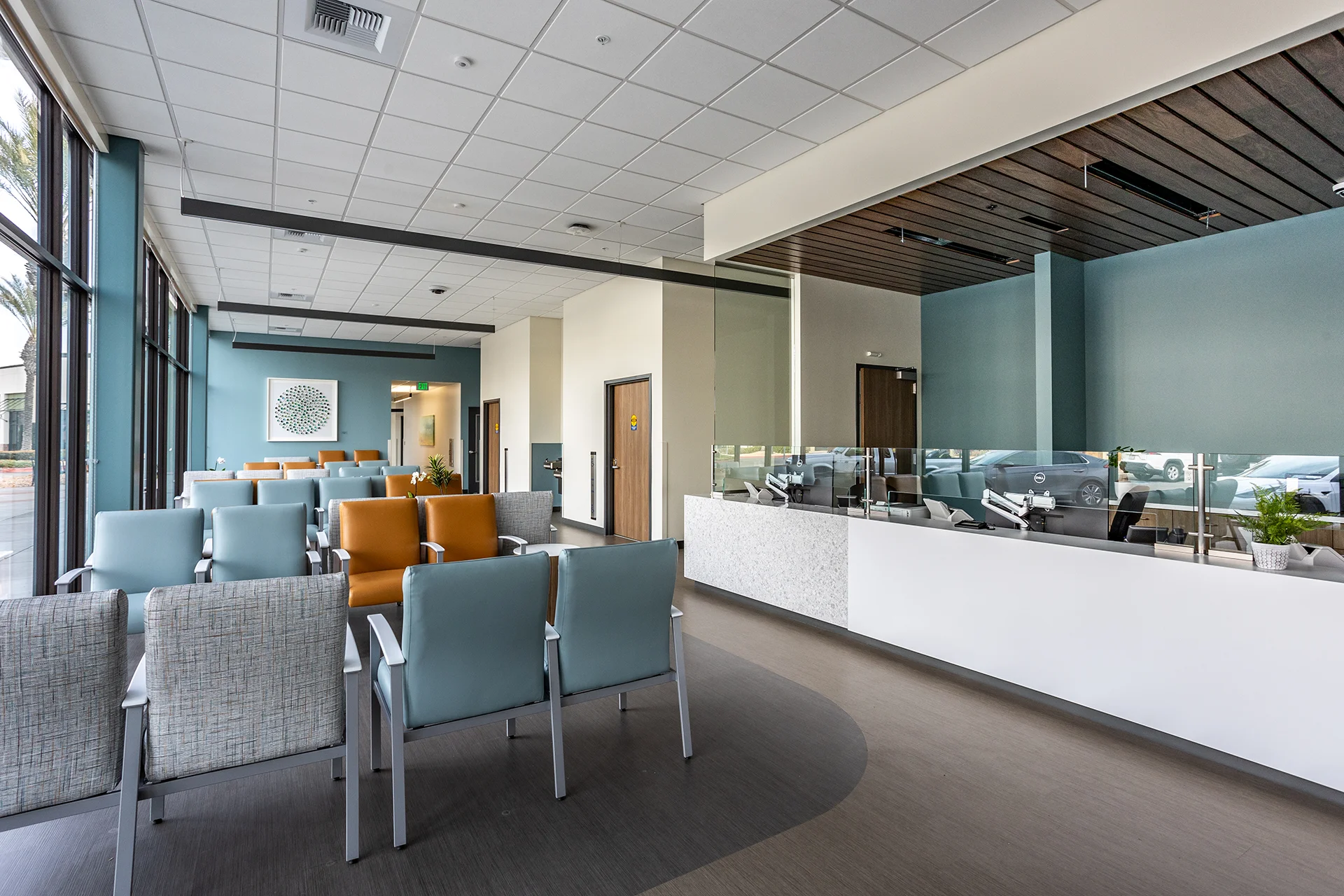 Comprehensive view of a medical facility entrance lobby in San Joaquin County, showcasing rows of blue and tan vinyl modular armchairs, patterned fabric accent seating, a white terrazzo multi-station check-in counter with glass privacy screens, and a dark wood slat ceiling cloud.