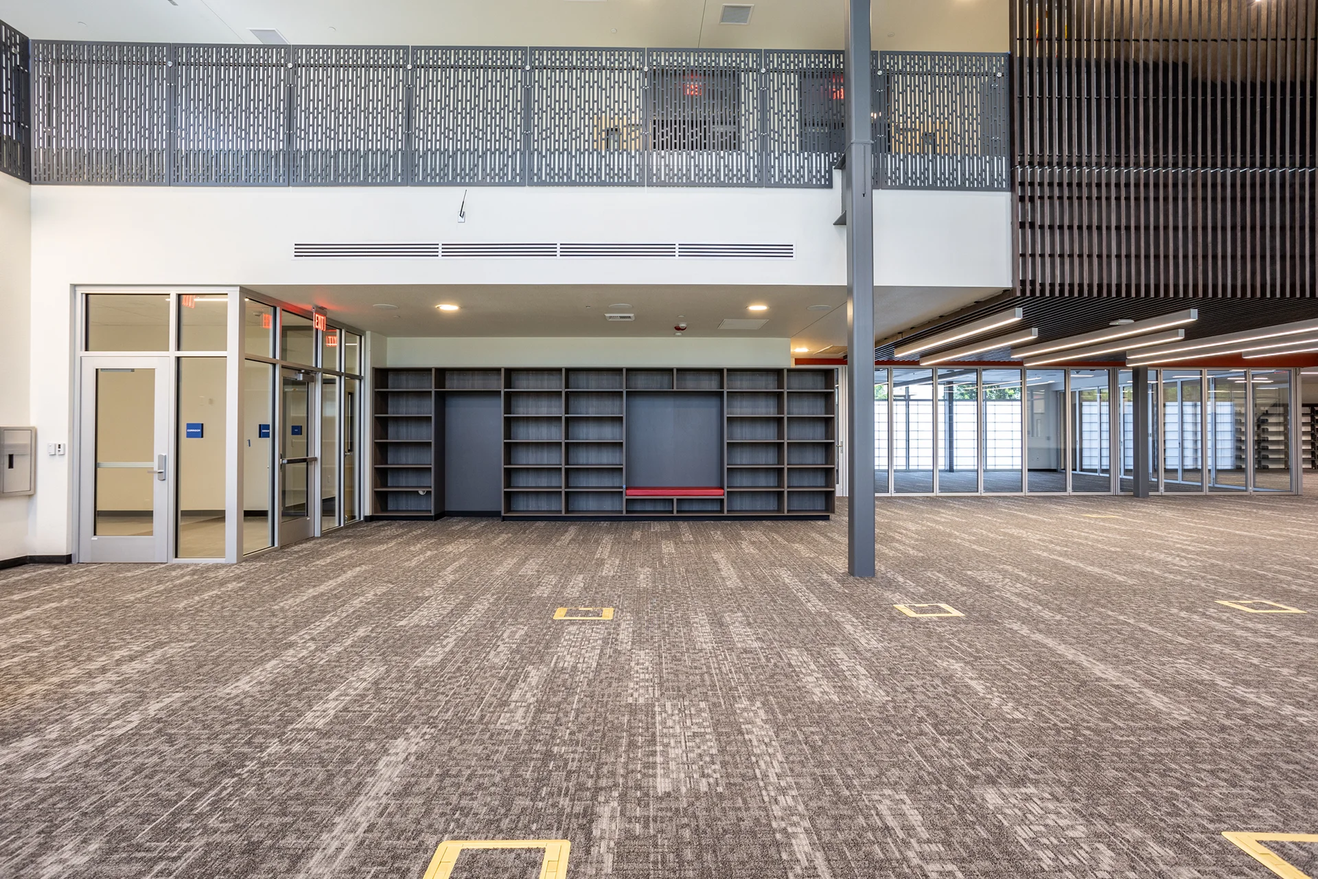 Academic campus multi-use learning space showcasing custom floor-to-ceiling dark wood shelving units with integrated cushioned bench seating, structural steel pillars, linear acoustic ceiling baffle details, and perimeter glass doors.