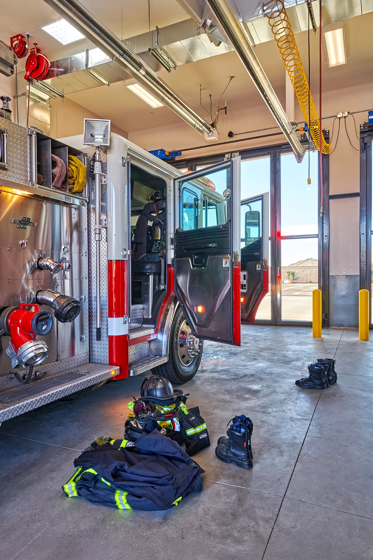 Side view of a parked fire apparatus vehicle with open passenger doors, showcasing integrated steel storage compartments and firefighter turnout gear including jackets, boots, and a helmet laid out on the bay floor.