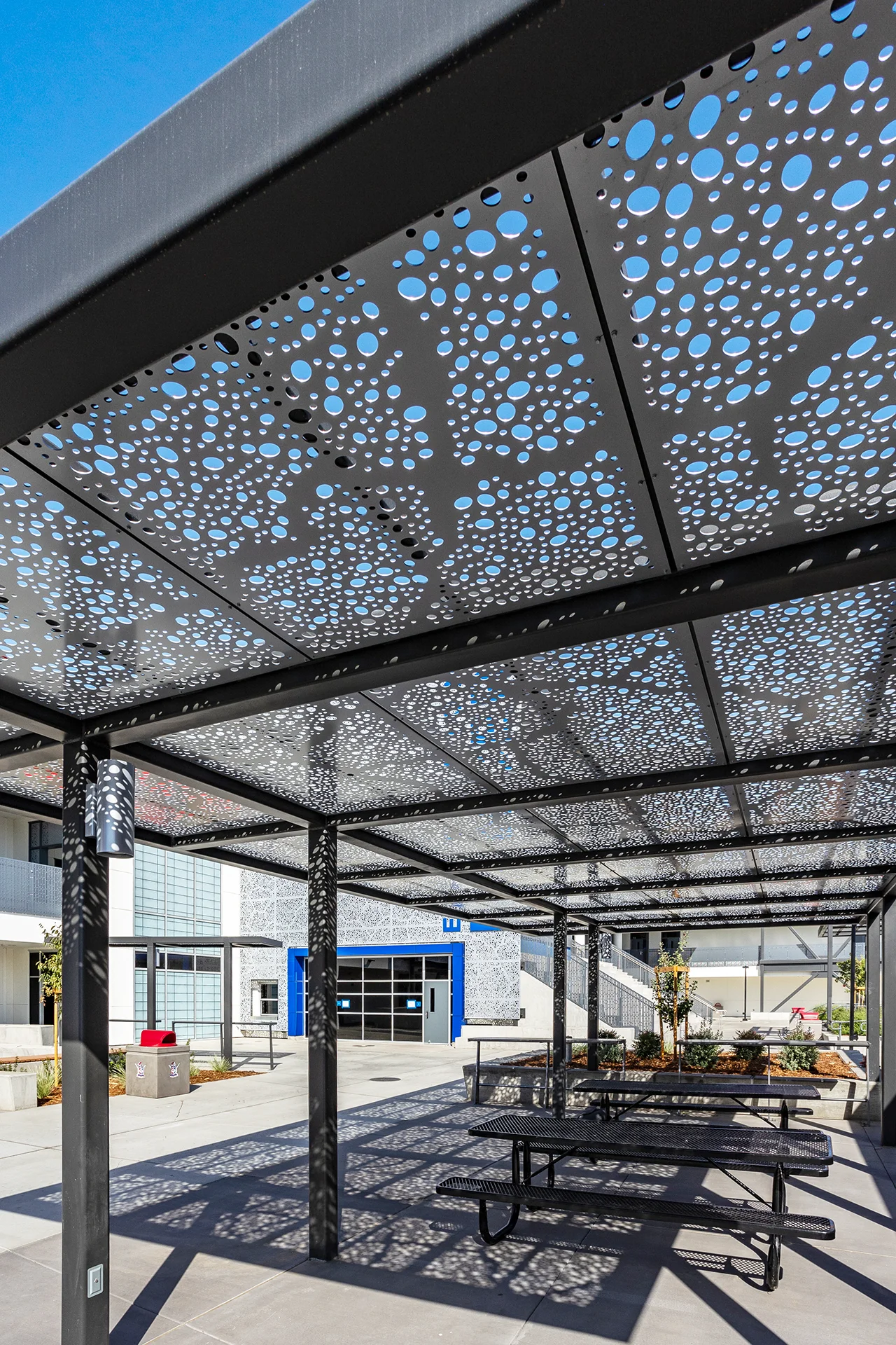 Close-up structural detail of a steel outdoor cafeteria pavilion frame fitted with custom perforated overhead panels that allow filtered sunlight through. The common area below contains heavy-duty black mesh steel picnic tables with integrated bench seating on a clean concrete pad.