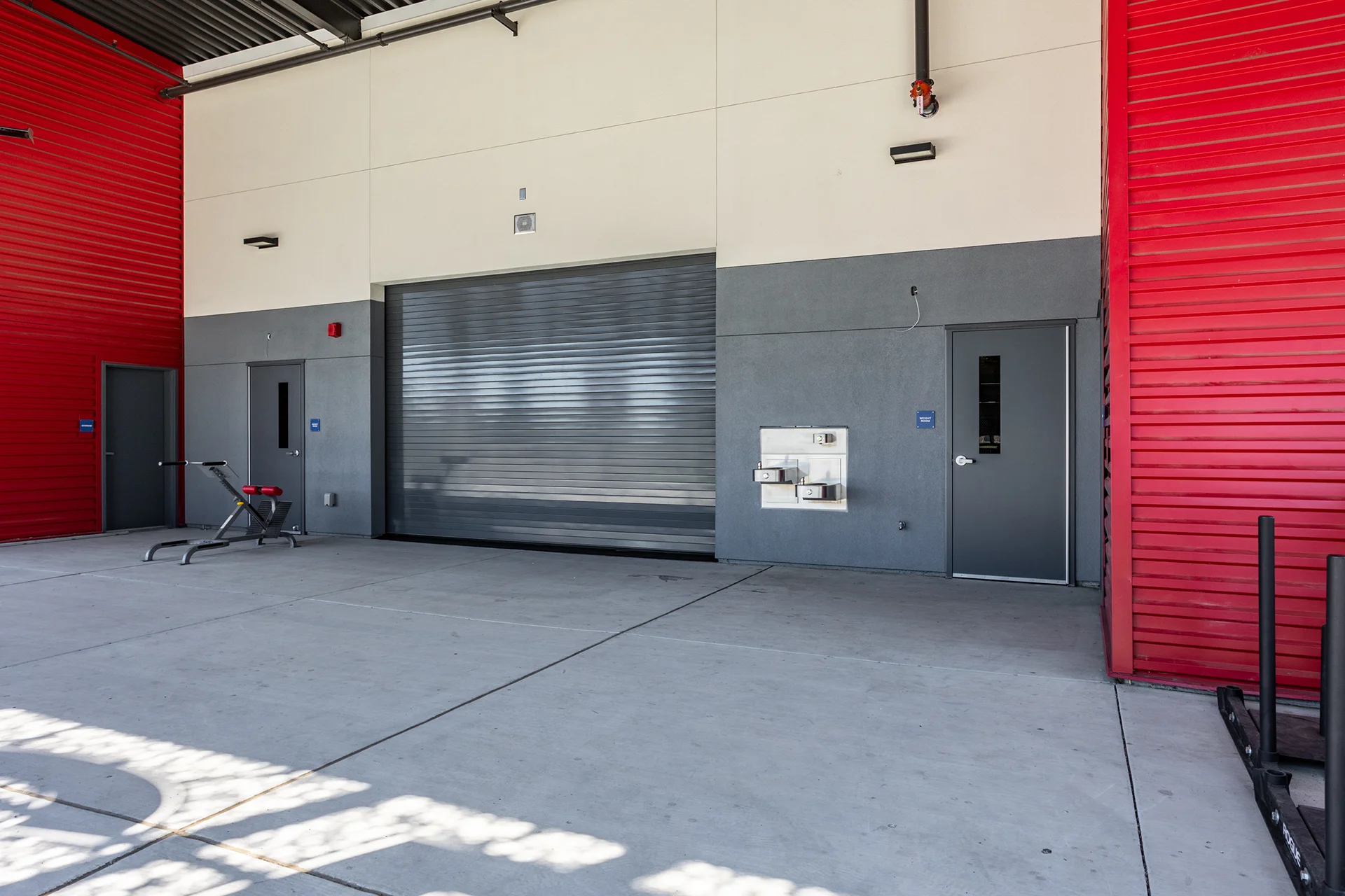 Exterior athletic training courtyard featuring cast-in-place concrete flooring, a gray metal roll-up garage service door, an exterior wall-mounted drinking fountain, and striking red corrugated metal siding accents.