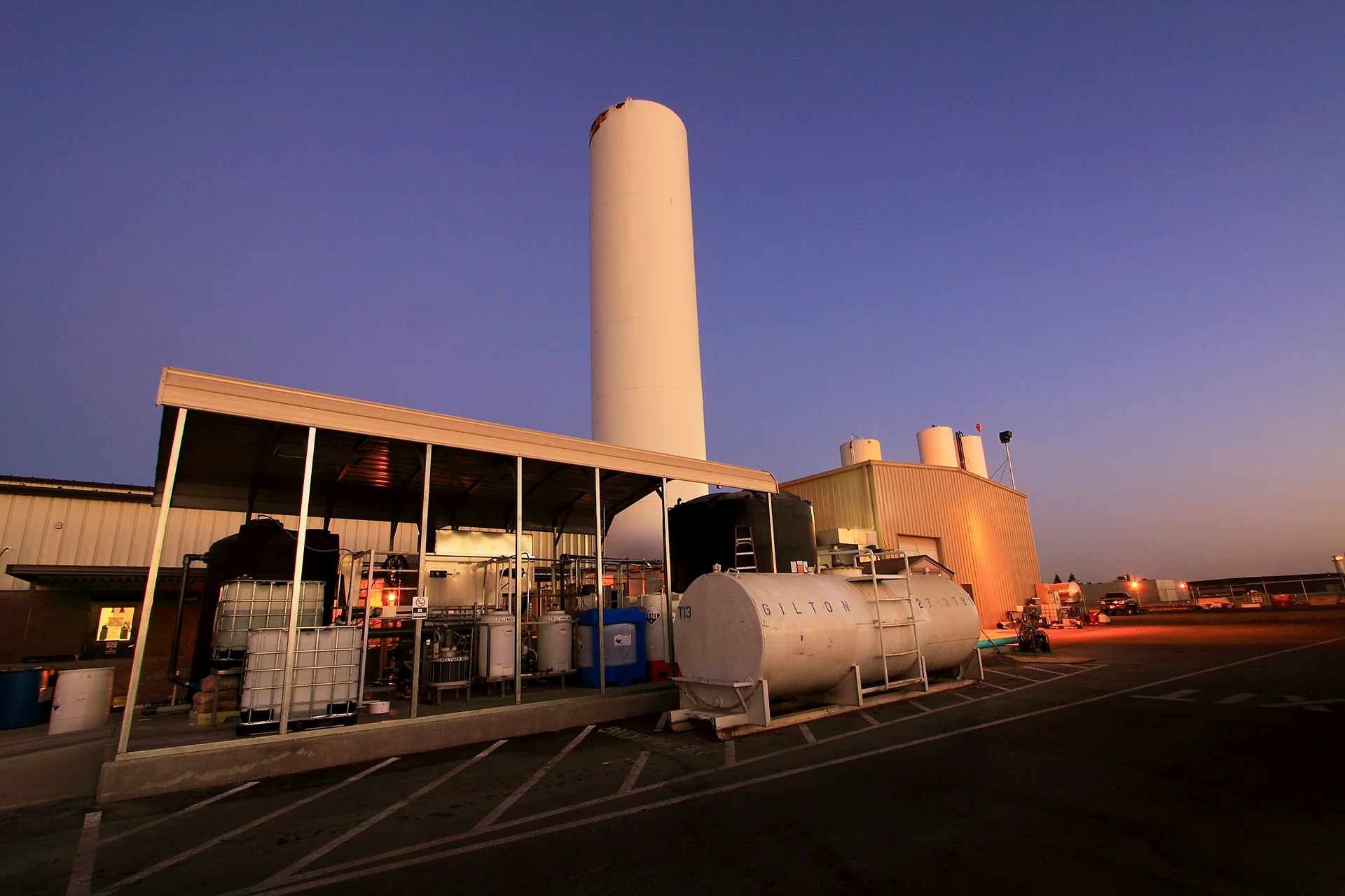 Low-angle shot of an outdoor utility area under a metal canopy structure, detailing commercial chemical storage totes, automated filtration equipment, an adjacent horizontal steel utility tank, a massive white storage silo, and asphalt paving with striped parking markings.