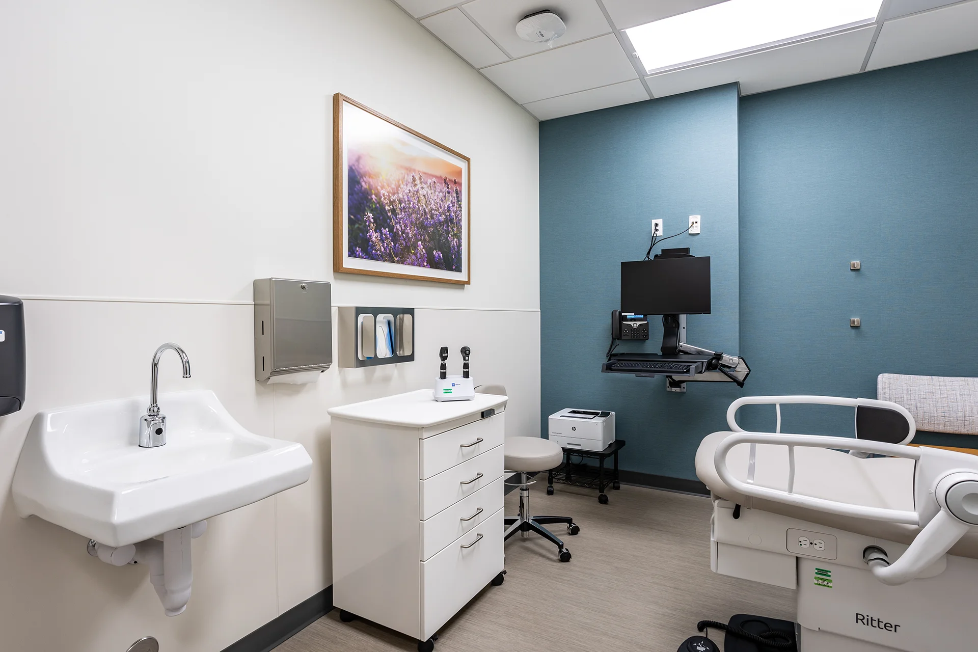 Well-lit clinical patient room showcasing a wall-hung vitreous china handwashing sink, a custom multi-drawer supply cabinet, a mobile doctor's stool, a wall-mounted co
