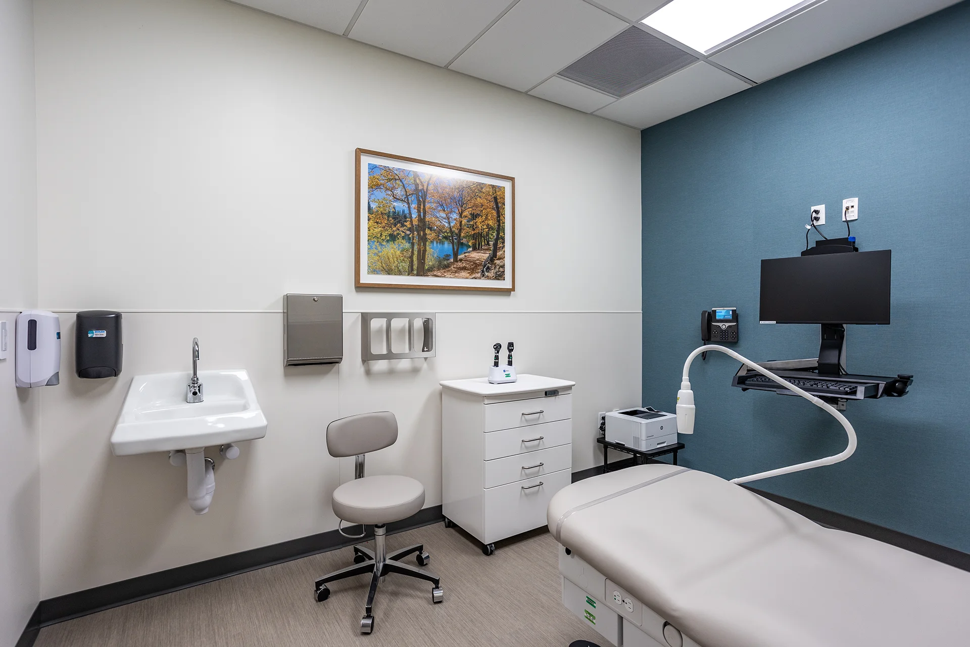 Standard medical check-up room displaying an adjustable bariatric exam bed with integrated paper roll, a white melamine supply cabinet with rolling casters, an ergonomic practitioner's stool, a wall-mounted vitreous china sink, and a deep teal blue vinyl-textured accent wall.