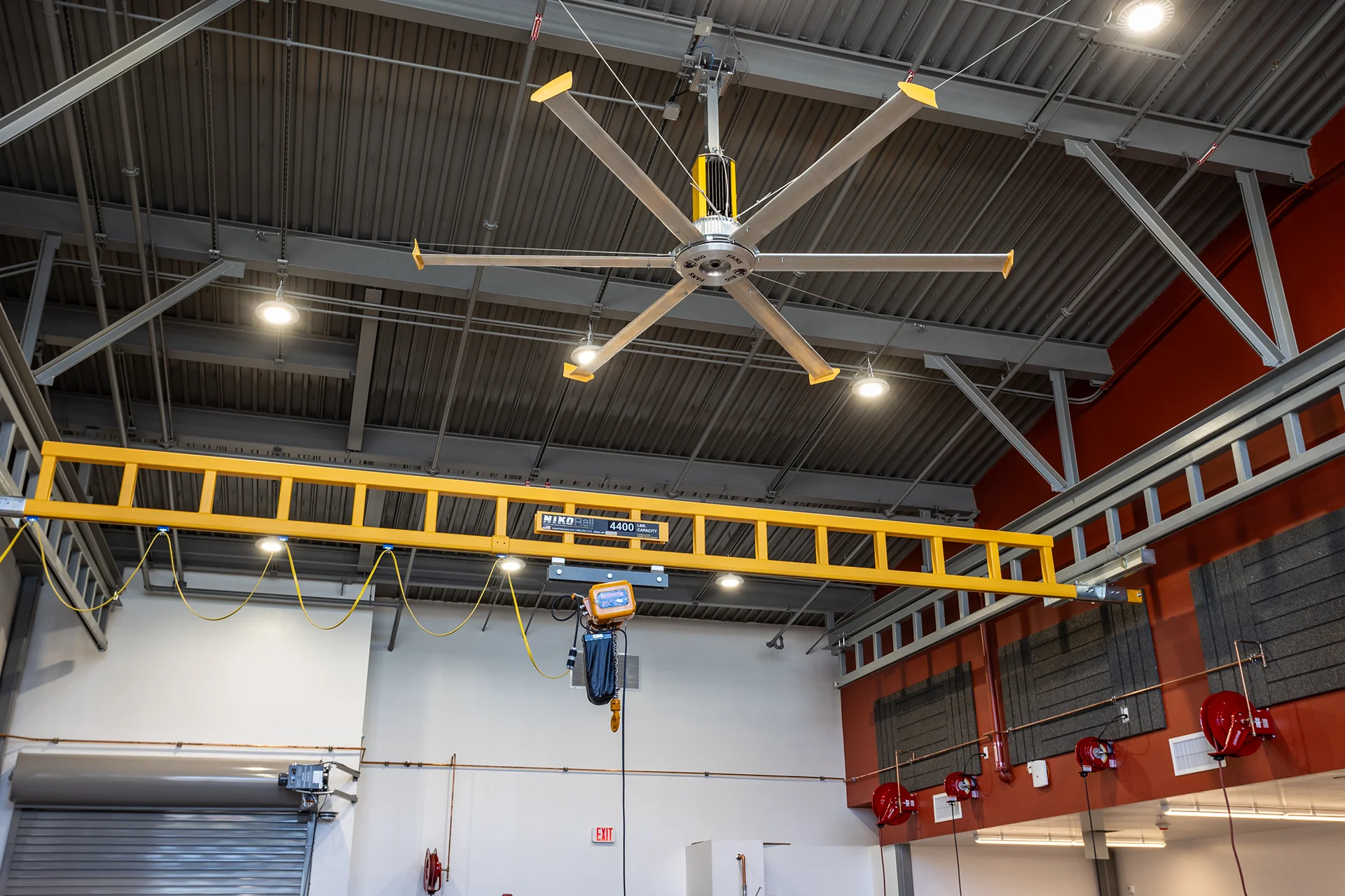Low-angle architectural view of an open-ceiling mechanical shop showing a high-capacity yellow steel bridge crane assembly with a rolling hoist mechanism, suspended beneath a multi-blade industrial HVLS ceiling fan and an exposed steel roof deck.