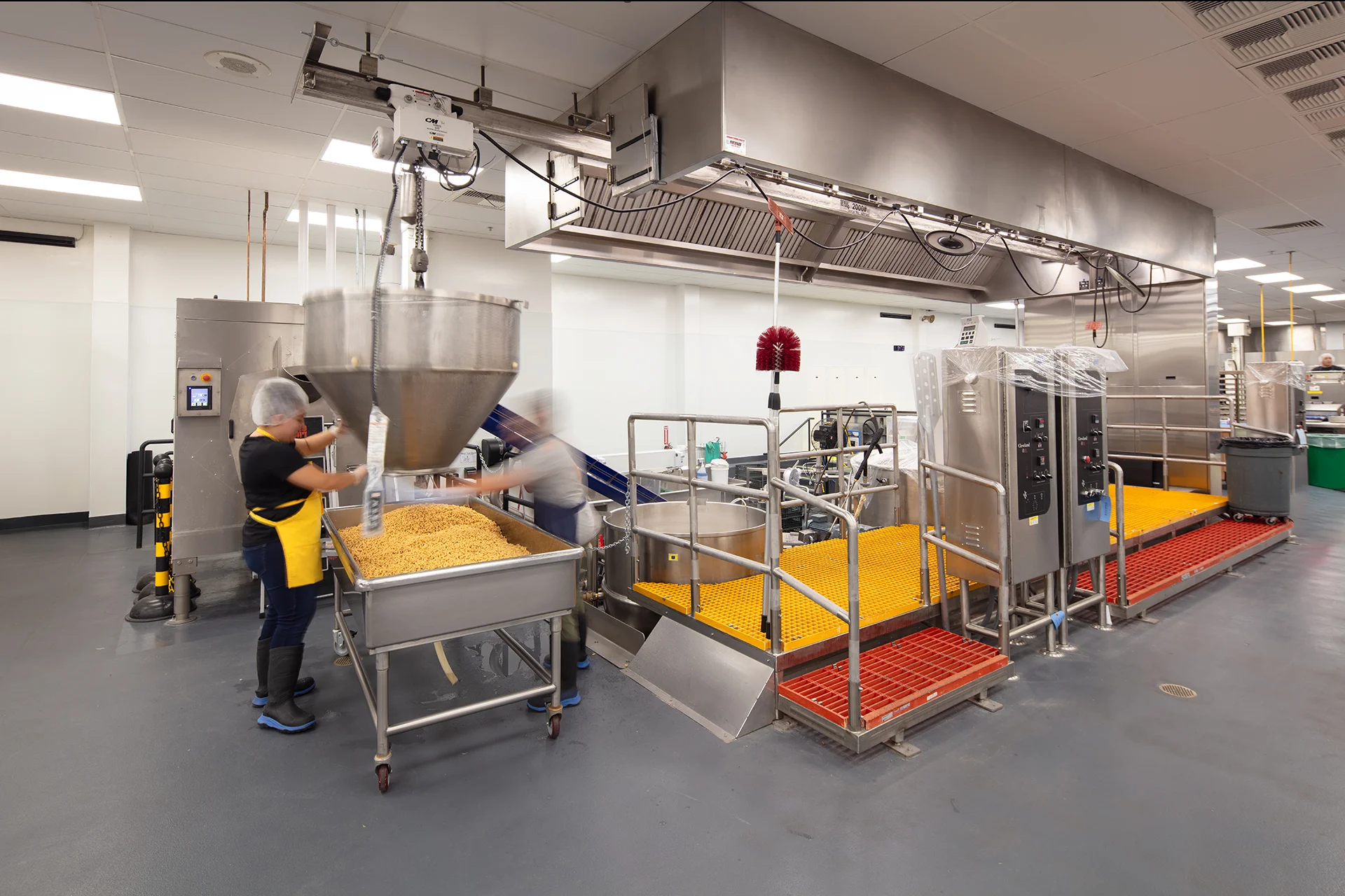 Industrial food-grade machinery installation highlighting an overhead mechanical hoist assembly, structural handrails, yellow safety platforms, and freestanding stainless steel electrical control enclosures.
