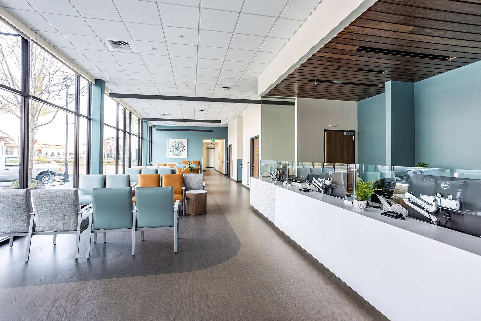 Bright healthcare clinic waiting room showcasing extensive floor-to-ceiling storefront glass windows, alternating rows of tan leather and blue vinyl medical lounge chairs, a white quartz check-in desk, and an acoustic tile drop ceiling with recessed linear accent lighting.