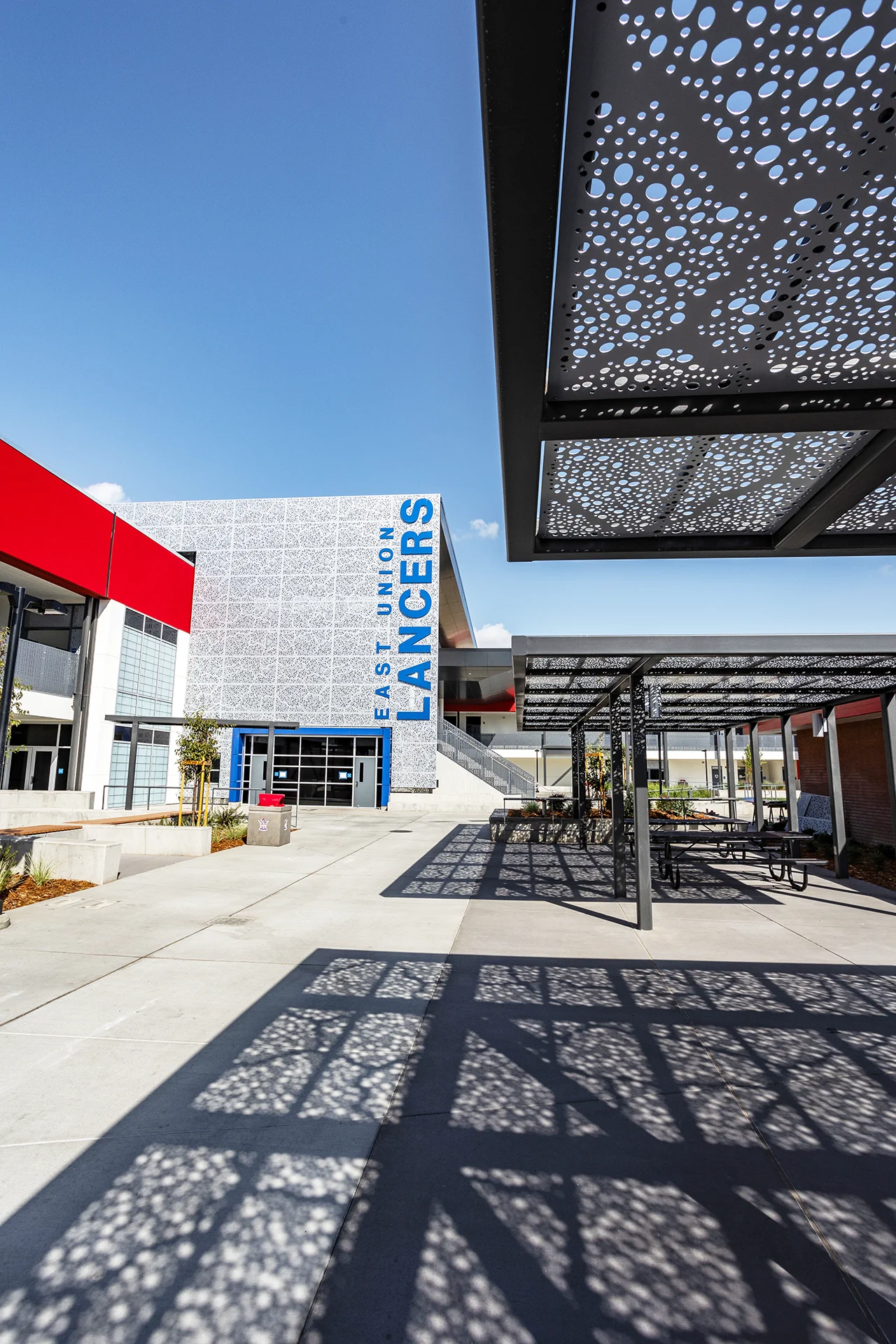 Upward perspective from beneath a black steel outdoor lunch pavilion showing the intricate shadow patterns cast by a perforated metal canopy. The background displays a modern high school building facade showcasing a textured white metal screen facade and prominent blue dimensional institutional lettering.