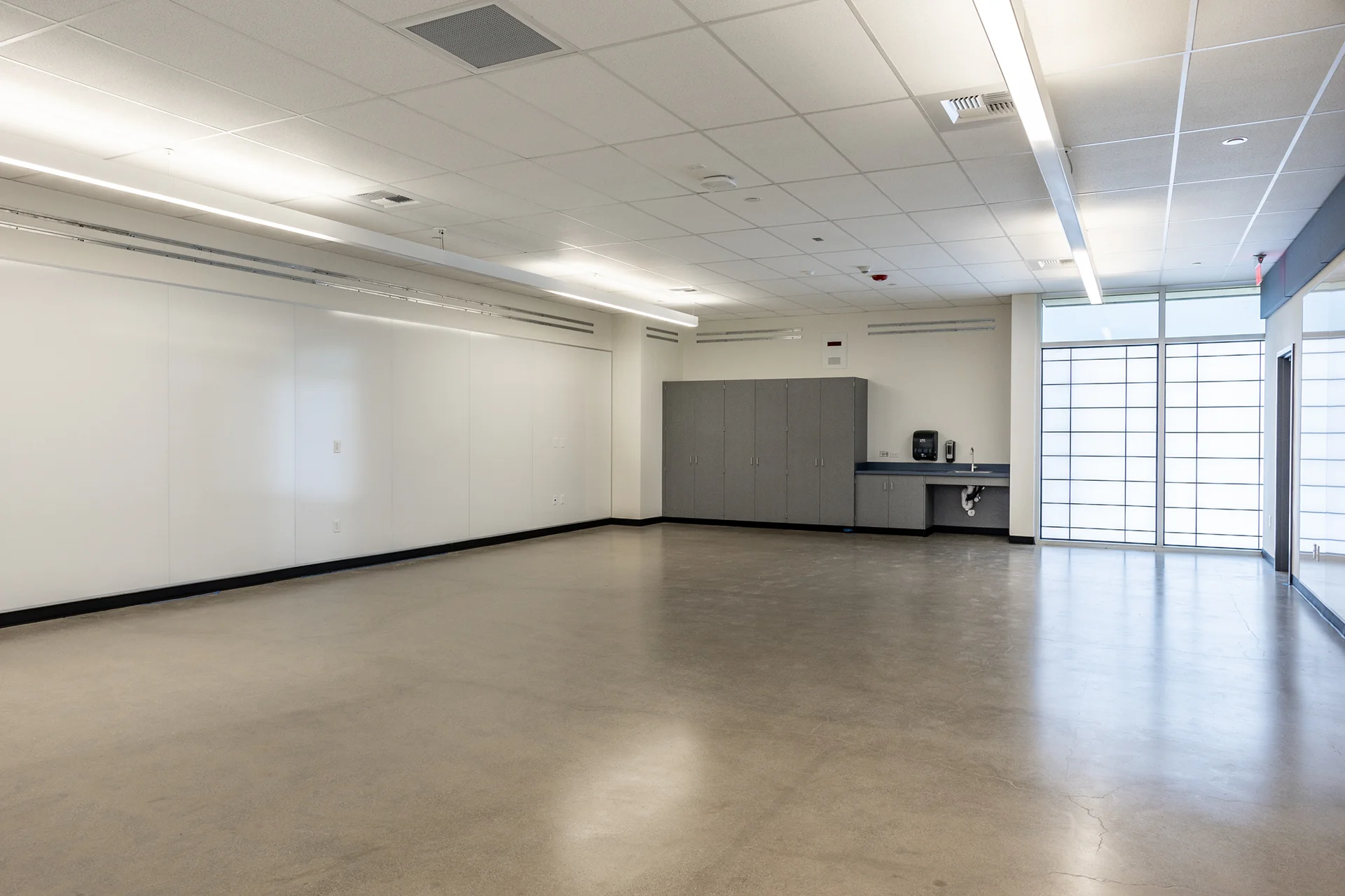 High school multi-purpose room showing polished concrete flooring, a long glossy white presentation wall, built-in gray counter cabinets with an integrated sink basin, and a gridded glass partition wall.