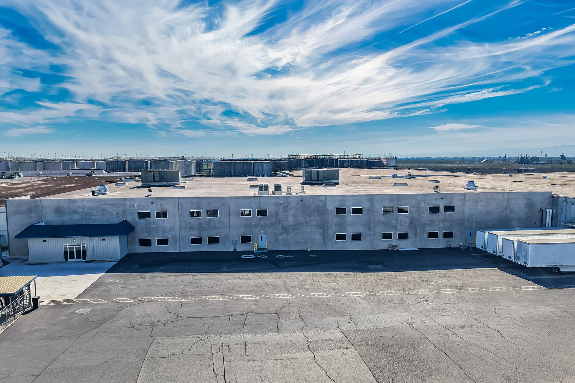Direct elevation drone view of a two-story tilt-up concrete industrial administrative facade, featuring architectural multi-pane windows, a structural entry vestibule with a blue metal awning, a cracked asphalt access driveway with safety cross-hatching, and industrial storage tanks along the horizon line.