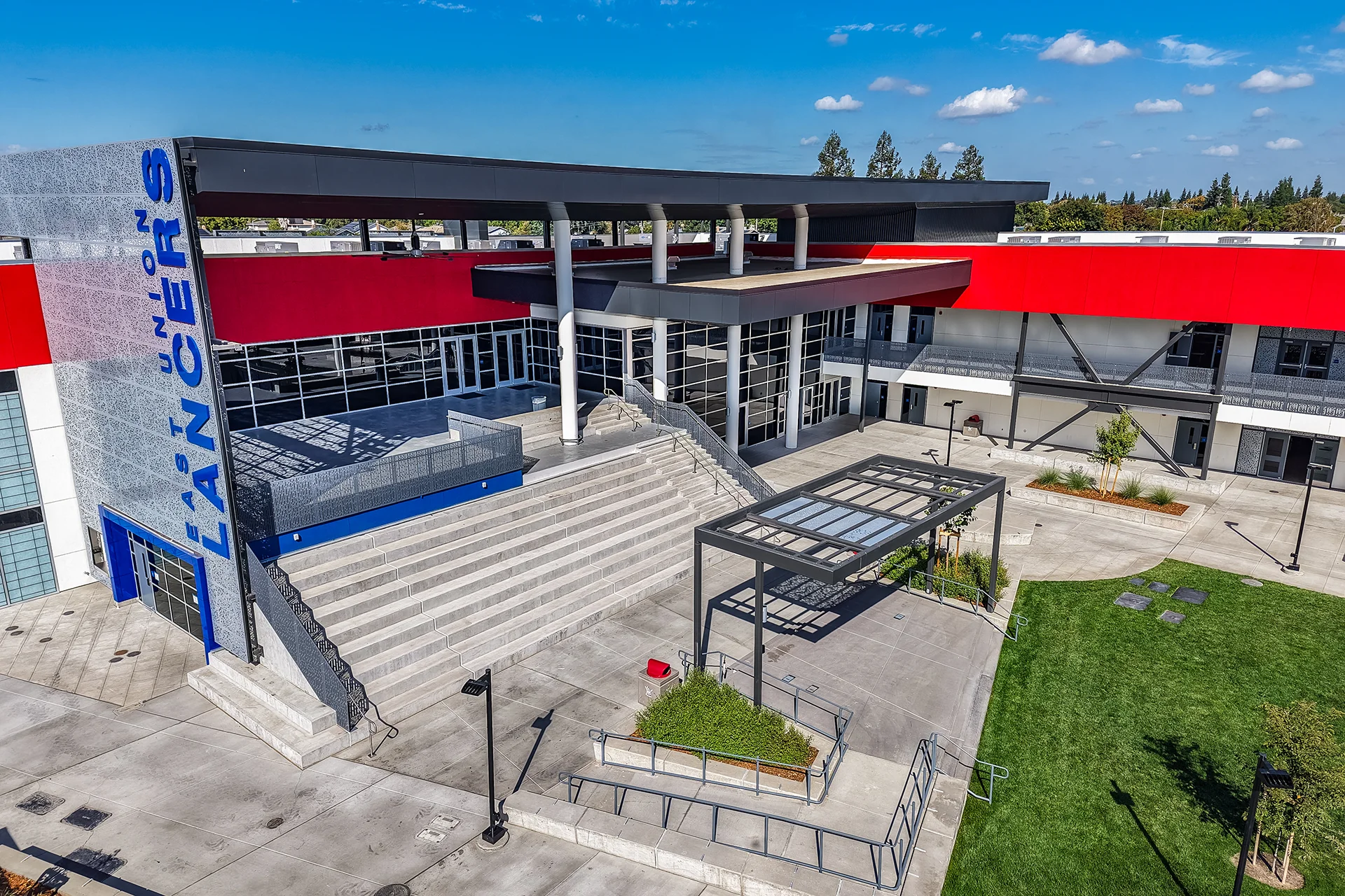 Close-up elevated view of a school courtyard presentation area highlighting a gray concrete grand staircase, defensive metal handrails, a blue perforated facade panel, a landscaped lower planter, and a central lawn panel.