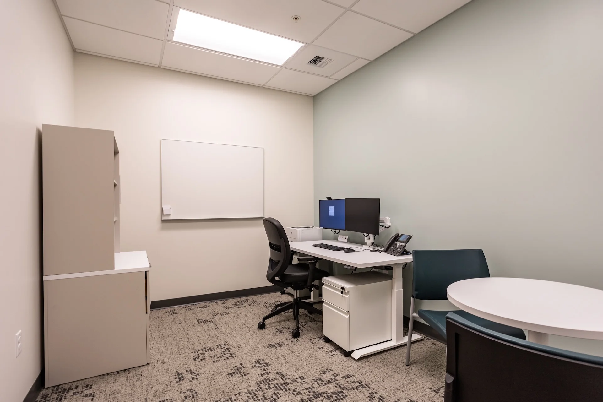 Shared administrative consultation room configured with an adjustable-height white desk, dual monitors, a separate round consultation table with fabric guest chairs, a magnetic wall-mounted whiteboard, and patterned low-pile commercial carpet tile.