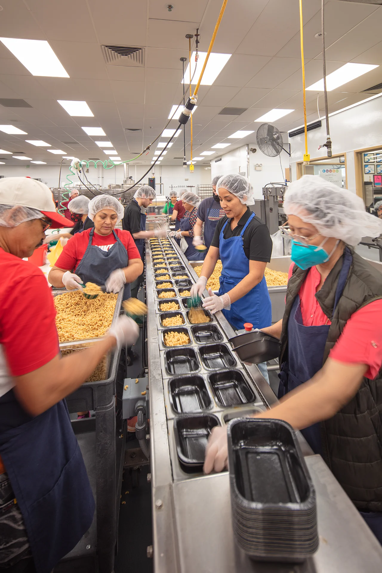 High-capacity food prep line configuration showing workers filling meal trays along an extended conveyor system with drop-down commercial utility lines and ceiling-mounted sanitation ventilation.