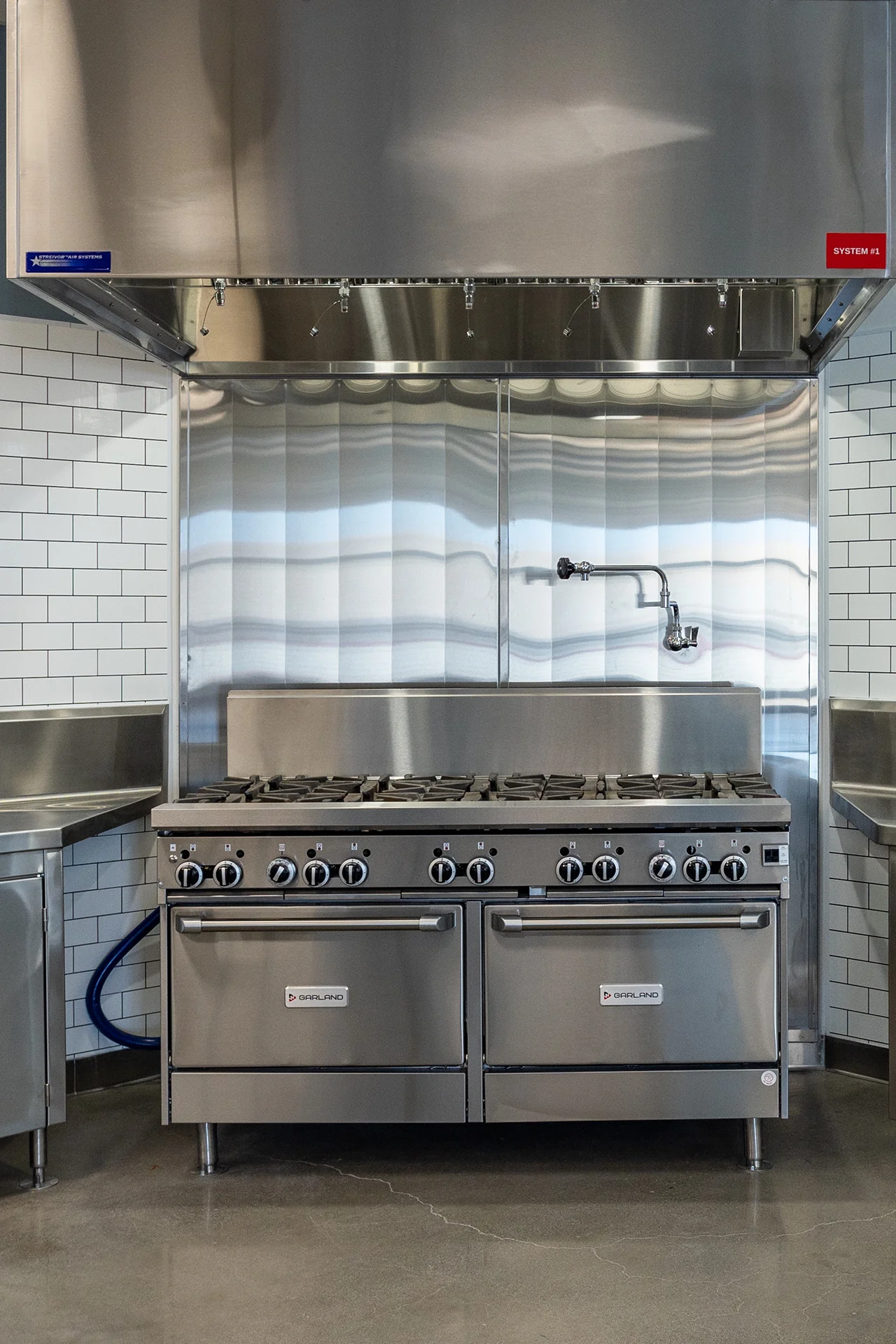 Heavy-duty commercial dual-oven gas range featuring cast-iron top grates and a articulated stainless steel pot-filler faucet, set against a polished metal backsplash panel under a fire-suppression hood.