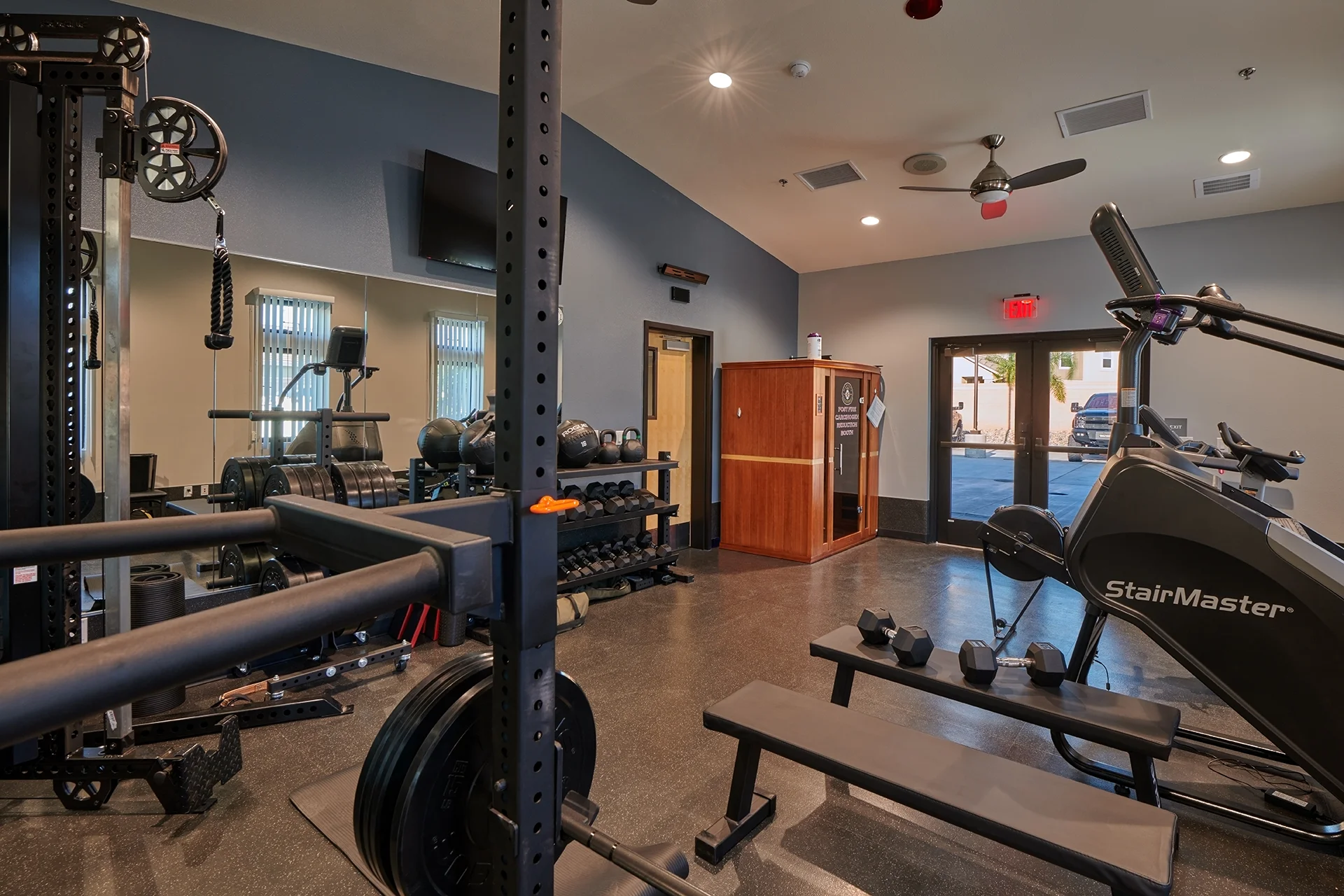 Physical training room highlighting a commercial steel weightlifting squat rack with barbell plates, wall-mounted mirrors, workout benches, and industrial rubber flooring under recessed ceiling lights.