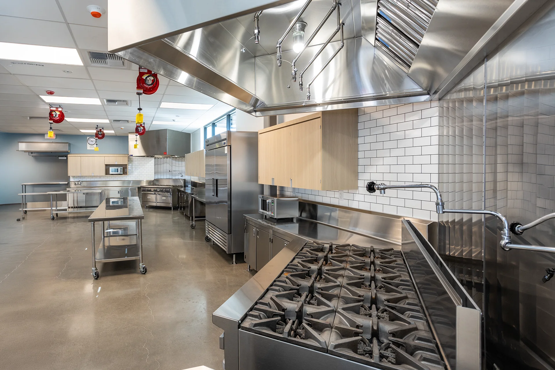 Angled view across a culinary education suite highlighting commercial gas ranges with stainless steel splash panels, overhead fire-suppression hoods with integrated lighting, reach-in coolers, and bright white subway tile walls.