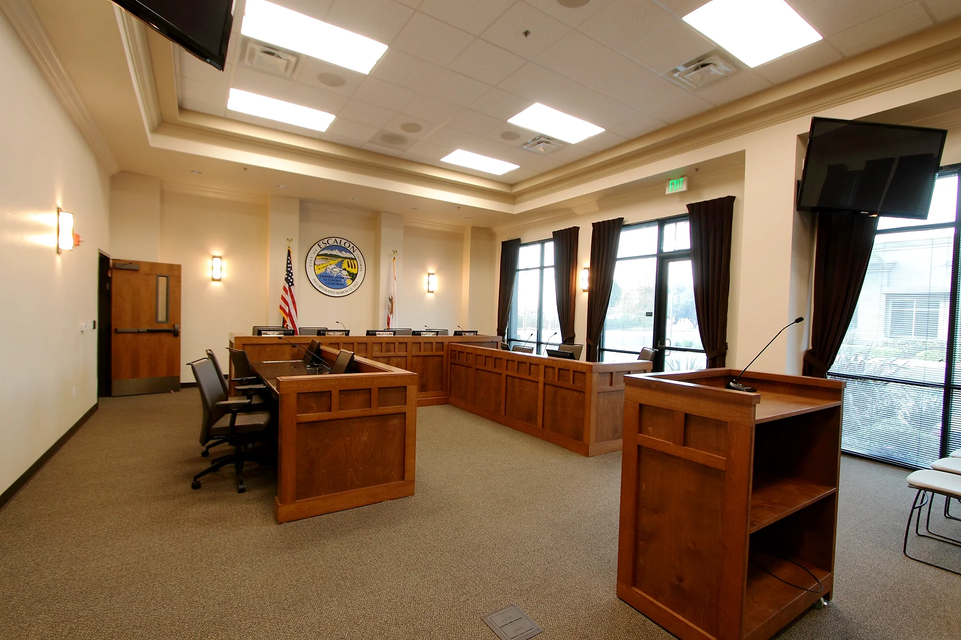 Front view of the municipal council chamber dais showcasing the City of Escalon circular wall seal, flanking American and California flags, a mobile wooden speaker lectern, and tall windows dressed with deep brown privacy drapery.
