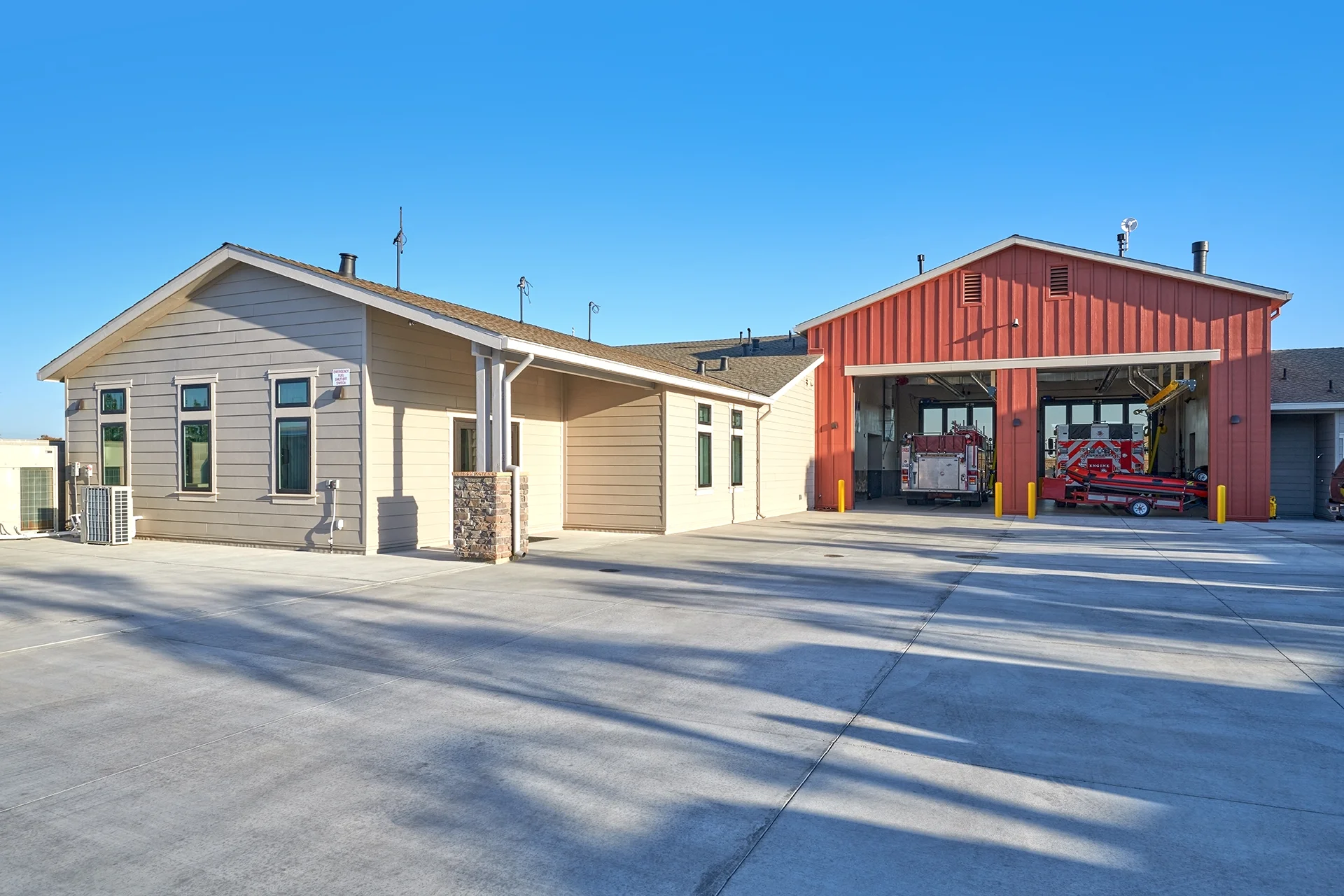 Rear perspective of the civic facility showing the large concrete turnout yard, the tan horizontal lap siding on the living quarters, and the red vertical board and batten siding on the active multi-bay engine garage.