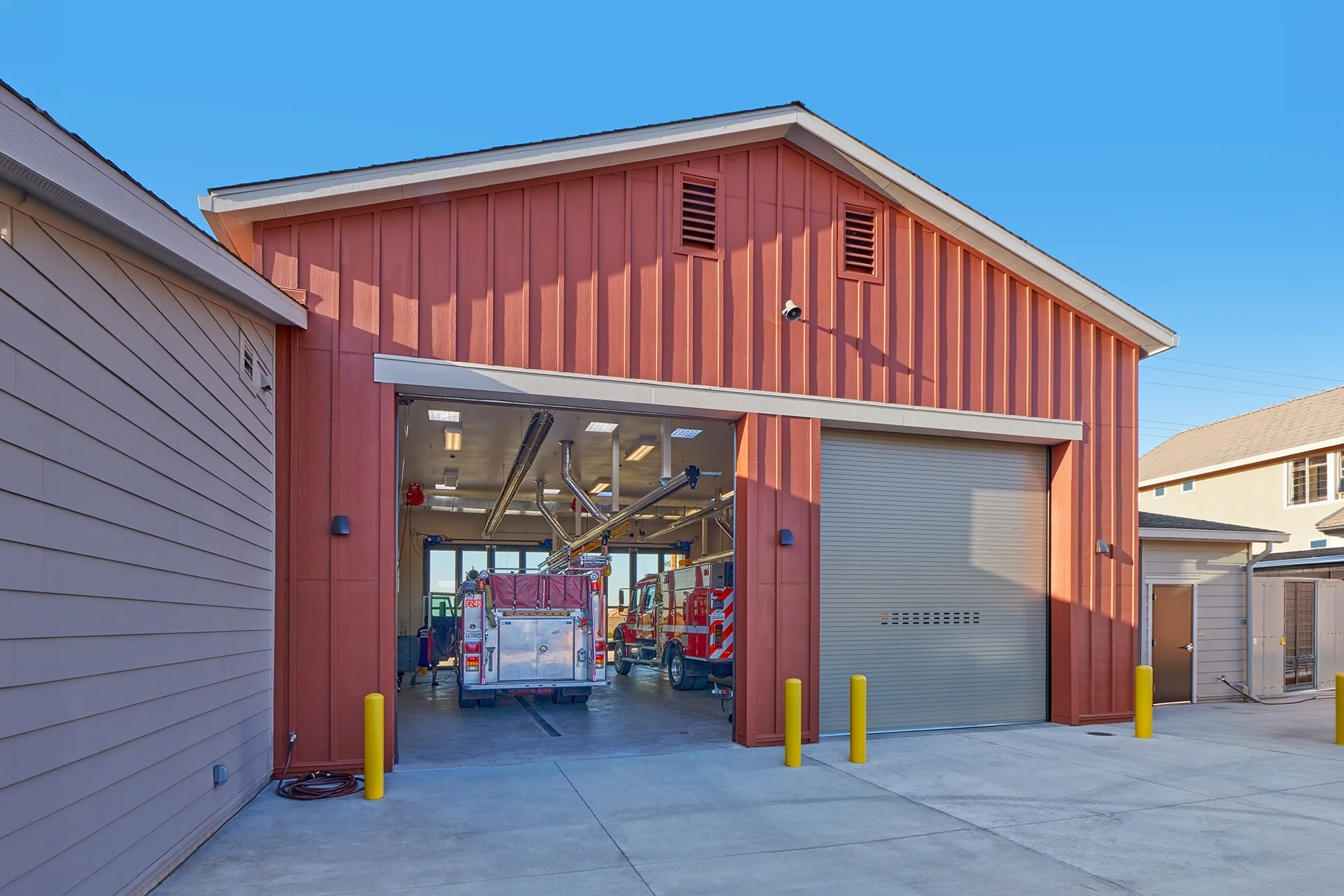 Rear elevation of the fire station apparatus garage showcasing red vertical board and batten siding, two large overhead rolling garage doors, concrete driveway, and yellow safety bollards under a clear blue sky.