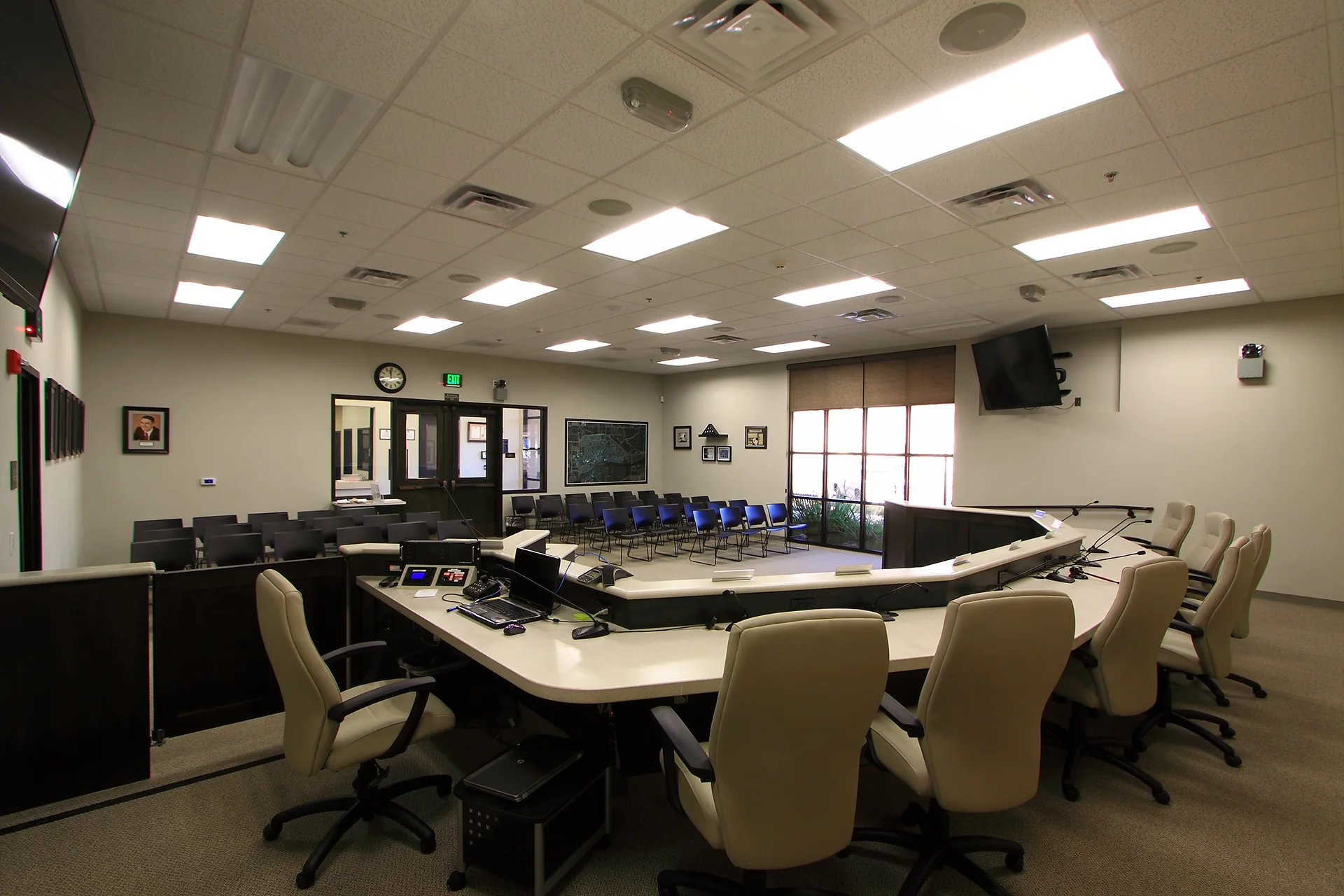 Interior view looking from behind the public official desk inside the council chambers, showcasing ergonomic tan swivel chairs, an integrated audio-visual system with computer monitors, a drywall drop-ceiling with recessed lighting grids, and rows of blue plastic visitor chairs.