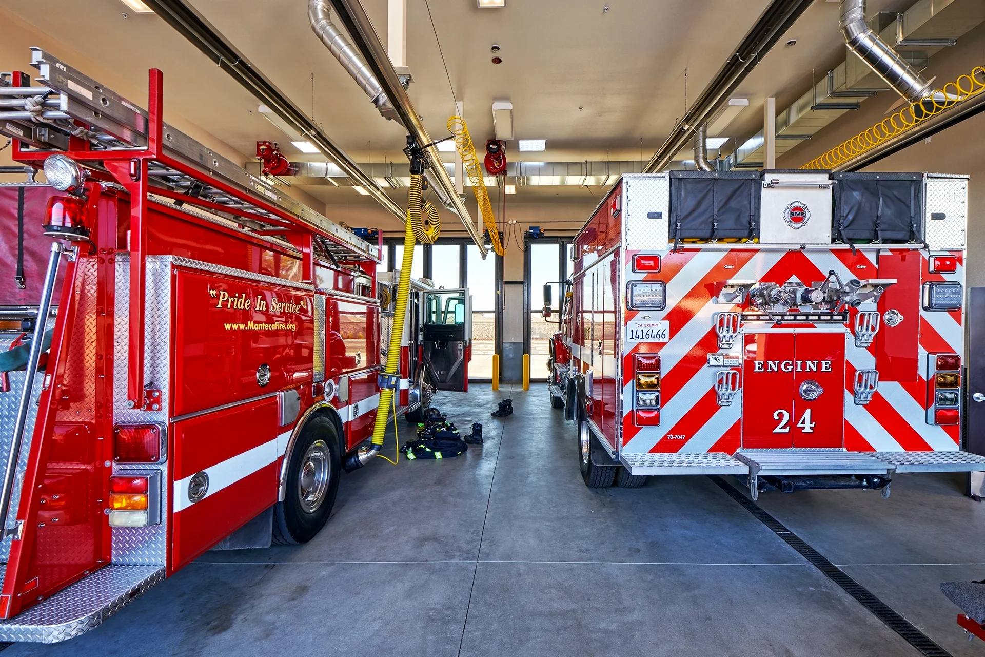 Wide view inside the open-ended apparatus bay housing multiple emergency vehicles, emphasizing the chevron reflective striping on the back of Engine 24, overhead utility conduits, and structural concrete flooring with drainage trenches.
