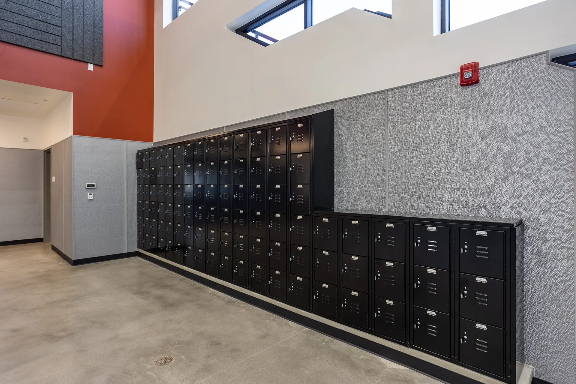 Institutional corridor featuring tiered banks of heavy-duty black steel lockers recessed against textured gray protective wall wainscoting, situated under high high-clerestory windows and an orange accent ceiling bulkhead with commercial fire safety devices.