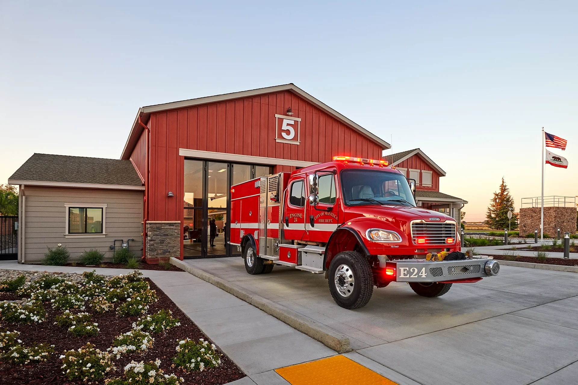 Action shot of a red commercial emergency vehicle with glowing red roof lights pulling onto the concrete access driveway from the station garage bay during twilight.