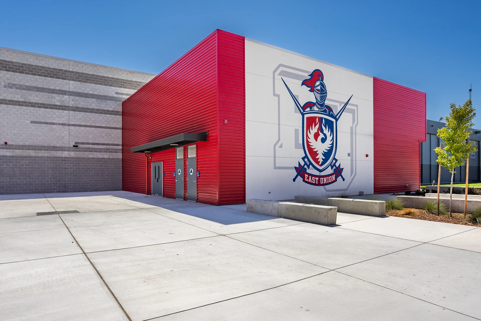 Exterior corner of an institutional school facility building showcasing a large painted knight mascot shield logo on smooth white plaster, contrasting red metal siding, cast concrete benches, and clean flatwork sidewalks.