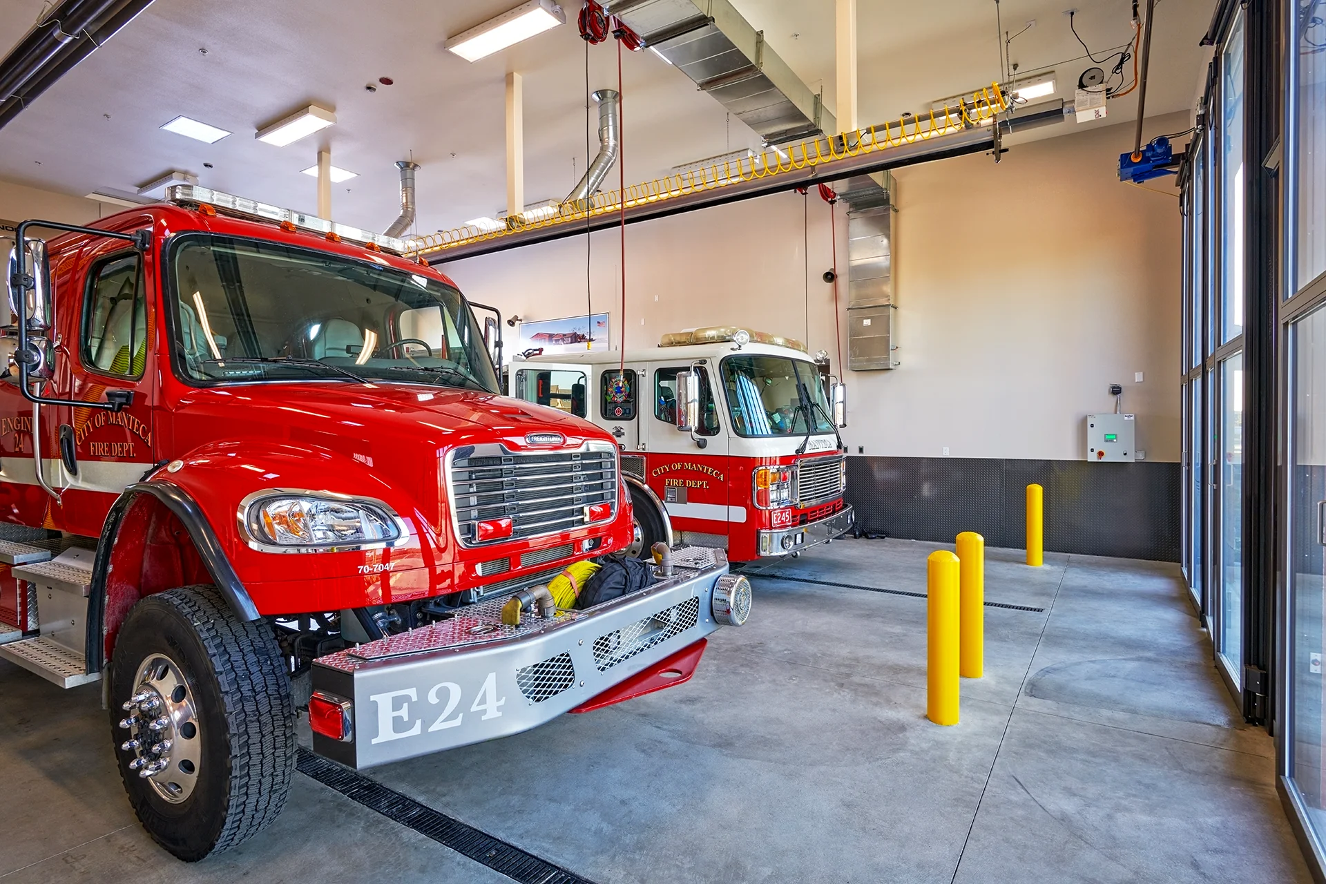 Close up front angle of emergency response vehicles including Engine 24 parked inside the municipal fire station bay, featuring overhead commercial utility lines, reflective security bollards, and full-height glass bifold doors.