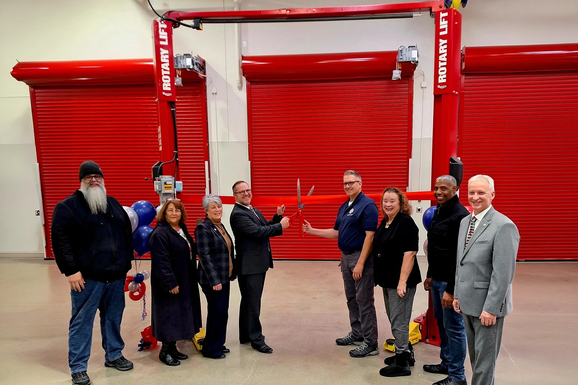 Community leaders and school officials gather for a ceremonial ribbon-cutting event inside a modern vocational training center, featuring commercial grade hydraulic vehicle lifts and red industrial roll-up warehouse bay doors.