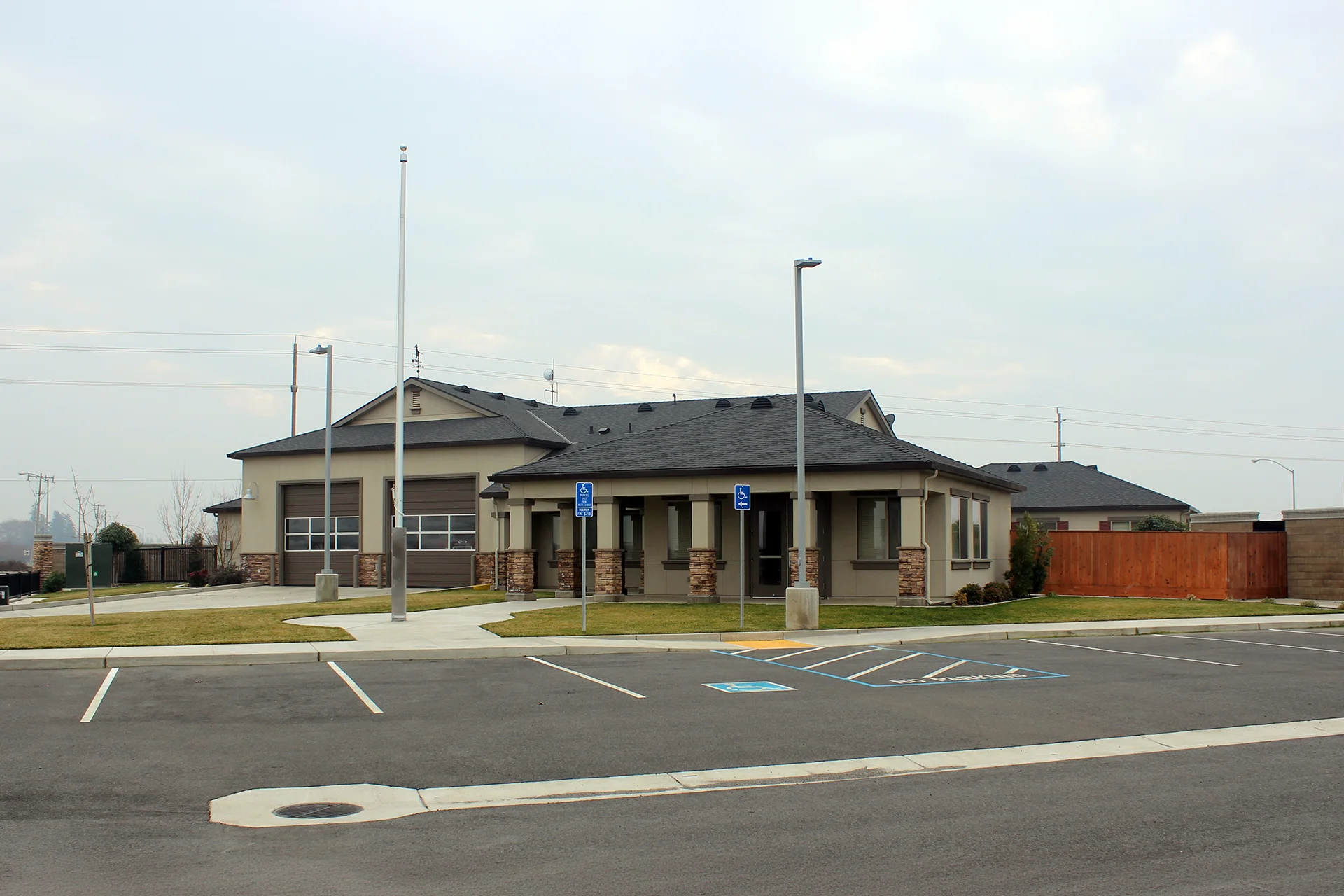 Exterior front perspective of the suburban Ripon Fire District Station 3 facility, highlighting the stone-veneer accent pillars across the office entryway porch, a paved asphalt parking lot with painted stalls, blue accessible parking signs, and dual garage bays.