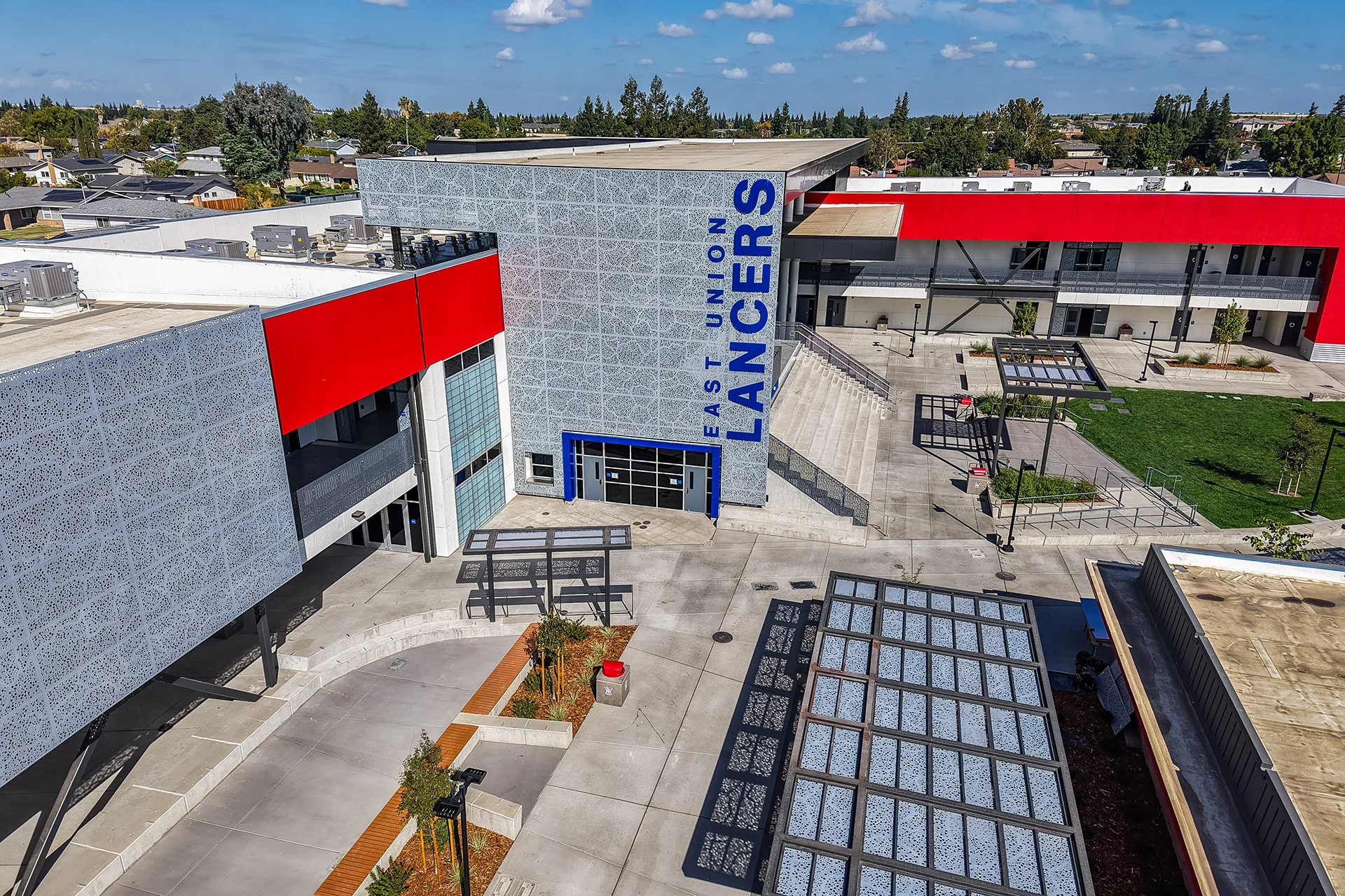 Roof-level aerial viewpoint of a commercial school facility modernization project showcasing gray structural canopies, multi-patterned concrete walking pads, architectural glazing, and vibrant red exterior finishes.