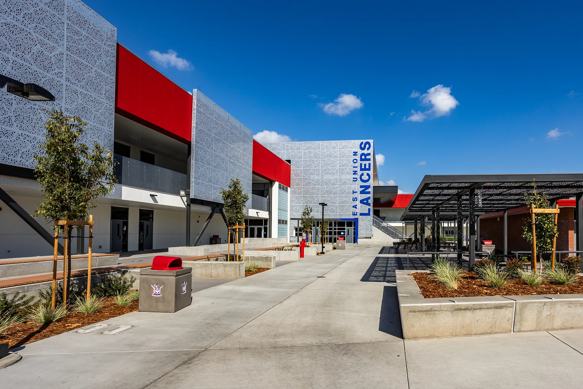 Wide perspective of the central quad at East Union High School, showing integrated concrete paths, landscaping planters with benches, a black steel trellis pavilion over picnic tables, and several multi-story school buildings with modern red and grey paneling under a blue sky.