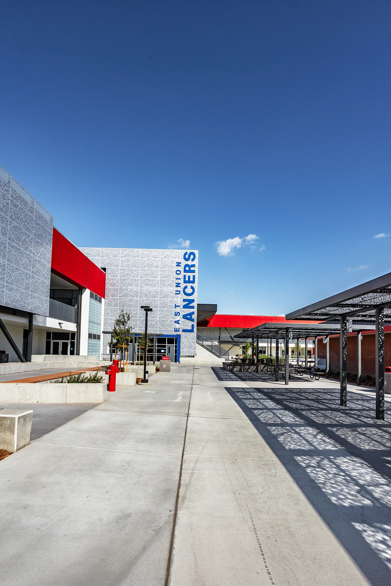 Wide campus walkway approach toward a striking architectural building wing featuring a perforated white patterned metal screen facade. The tower displays vertical blue dimensional block lettering that reads East Union Lancers above a storefront glass door entrance and a black steel lunch table pavilion.