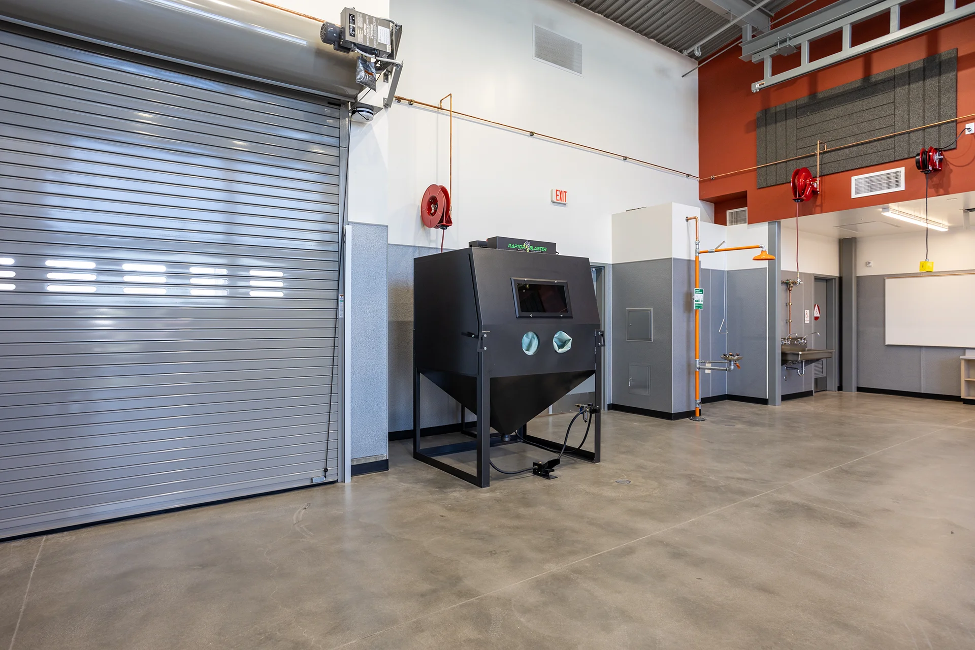 Wide-angle view across a technical education shop showcasing a large motorized metal coiling overhead door, a media blasting station, an emergency safety shower column with an eyewash bowl, and a wall-hung multi-faucet handwashing station.