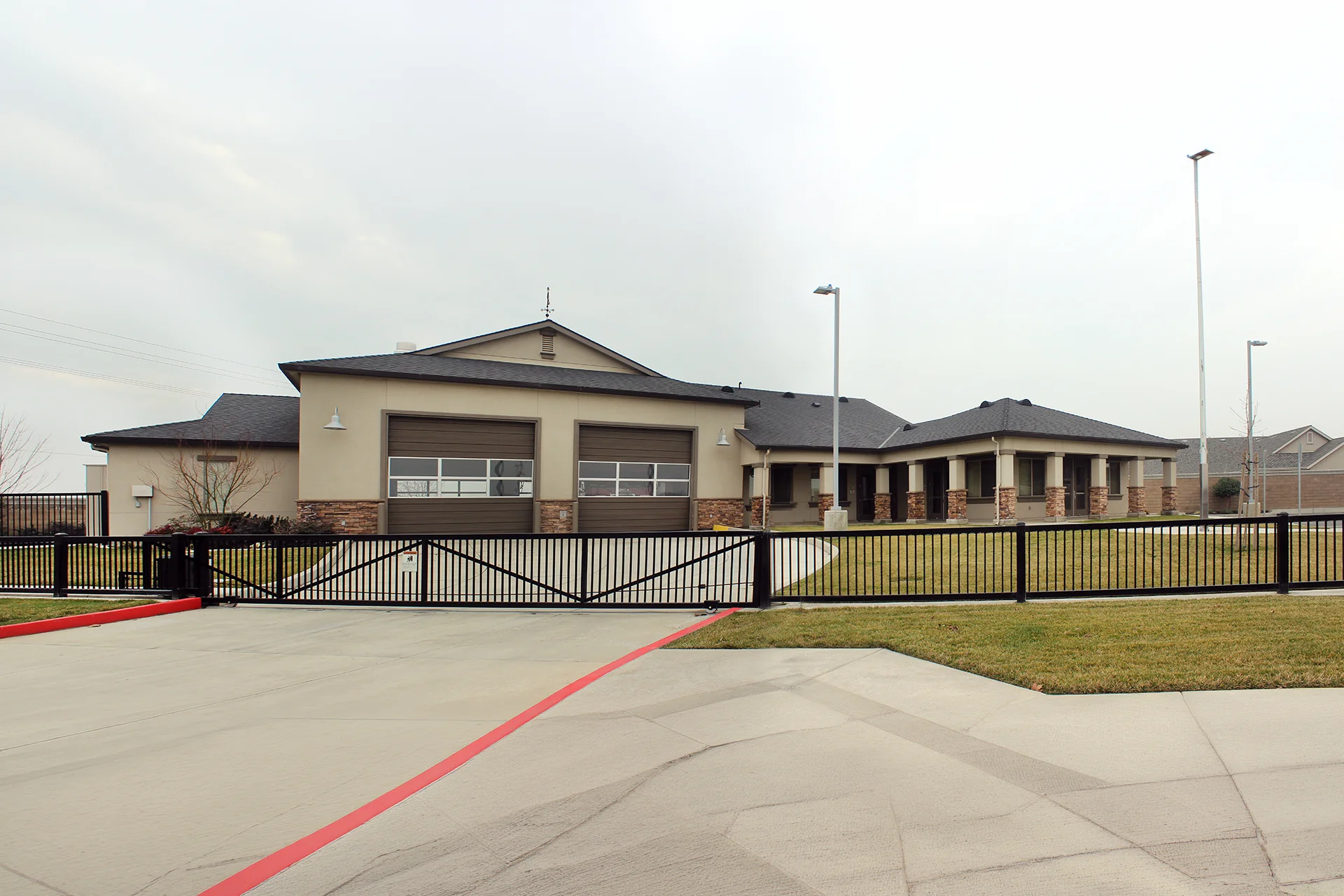 Ground level shot of the municipal facility focusing on a black motorized steel security gate system built across the concrete engine bay driveway, featuring dark bronze sectional overhead doors and a low-maintenance stucco exterior.