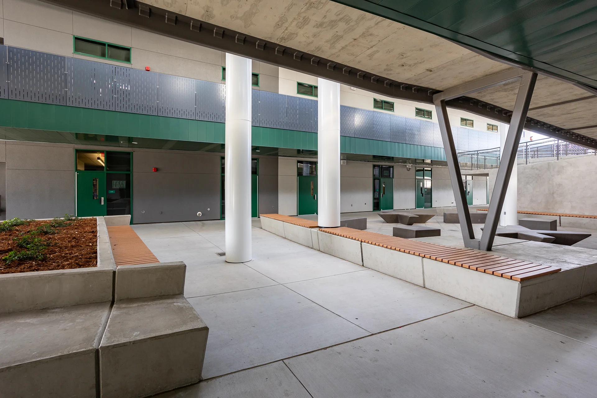 Ground-level perspective beneath a raw concrete overhang looking into a school courtyard plaza in Manteca, California, displaying long cast-in-place concrete planters topped with wood-slat bench seating, fresh bark mulch beds, structural steel V-bracing, and polished white columns.