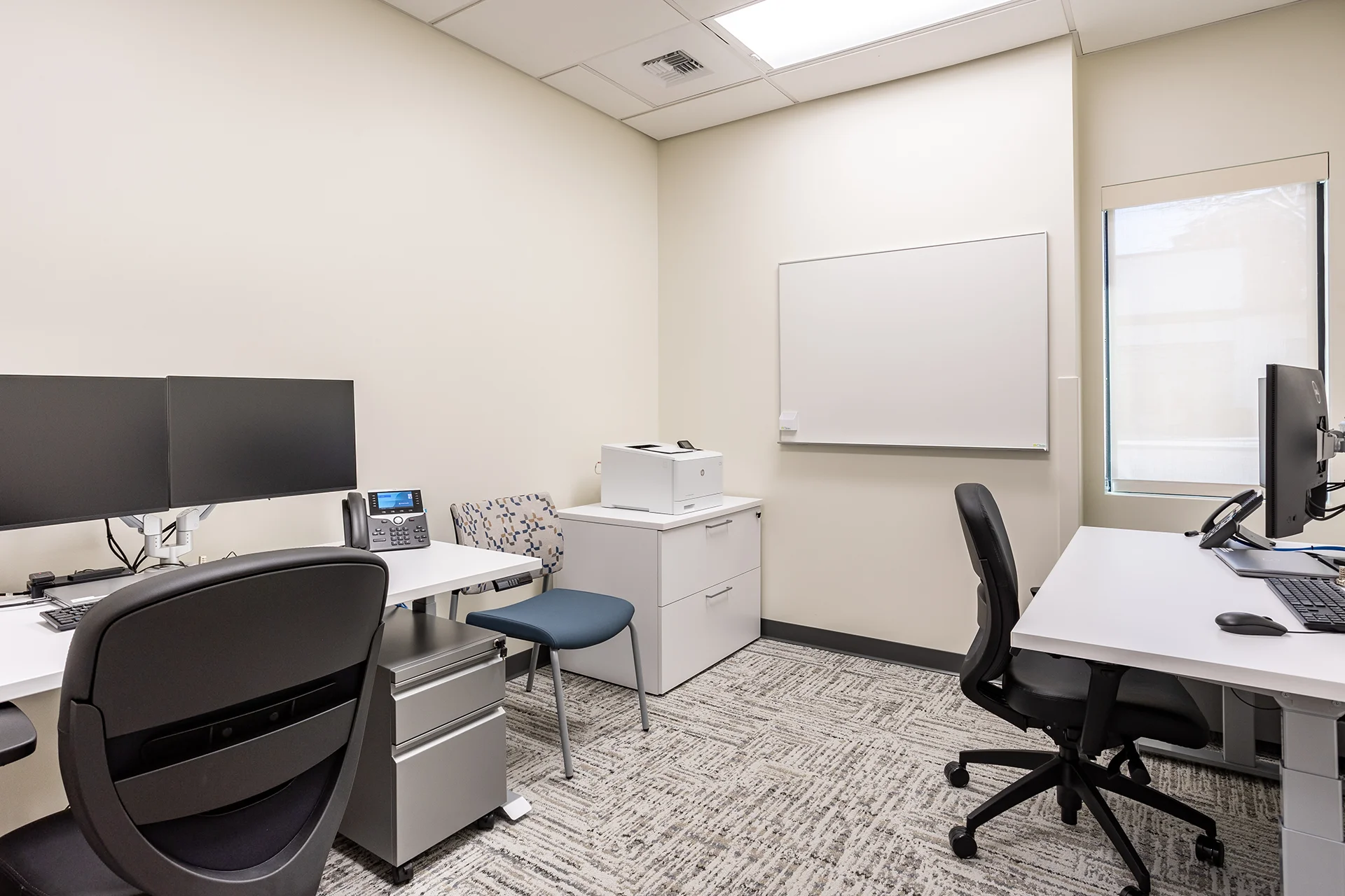 Dual-occupancy medical clinic office showing two adjustable height sit-stand desks, computer monitors, a wall-mounted porcelain whiteboard, a multi-drawer metal filing cabinet with an integrated desktop printer, and light grey commercial carpet tiles.