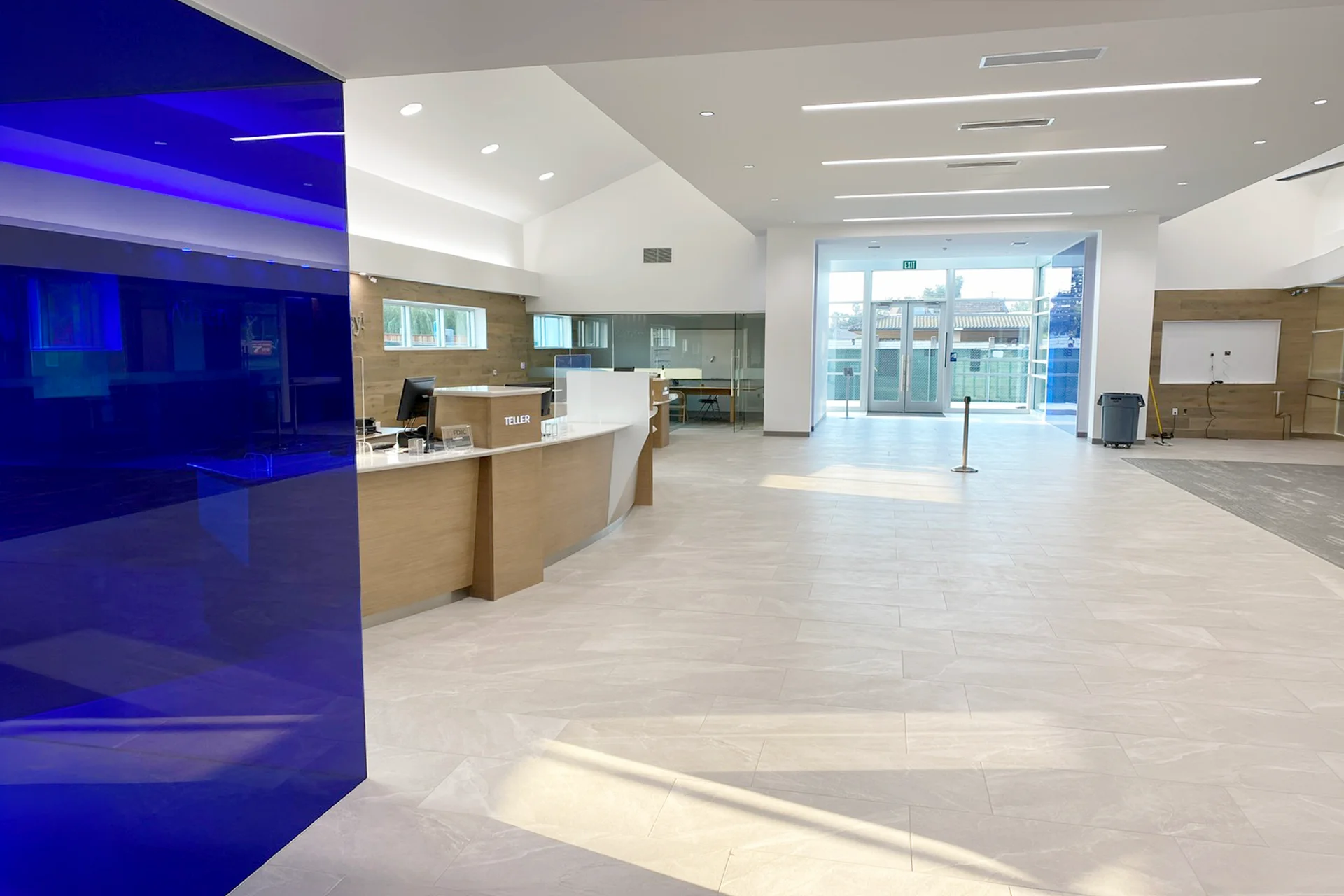 Interior perspective of the financial branch lobby looking toward the rear glass exit doors, highlighting a glossy, translucent blue glass partition wall in the foreground, recessed linear ceiling lighting, and built-in transaction counters.
