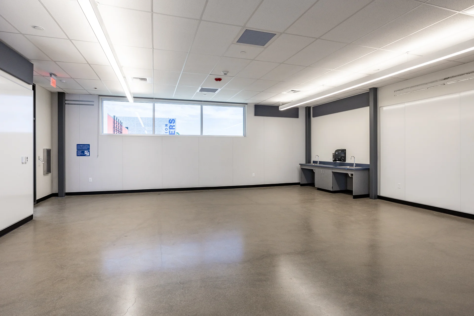 Renovated school classroom interior featuring a sealed, reflective concrete floor, minimalist white wall panels, structural steel pillars, and a dual-sink sanitation station beneath a wide window.