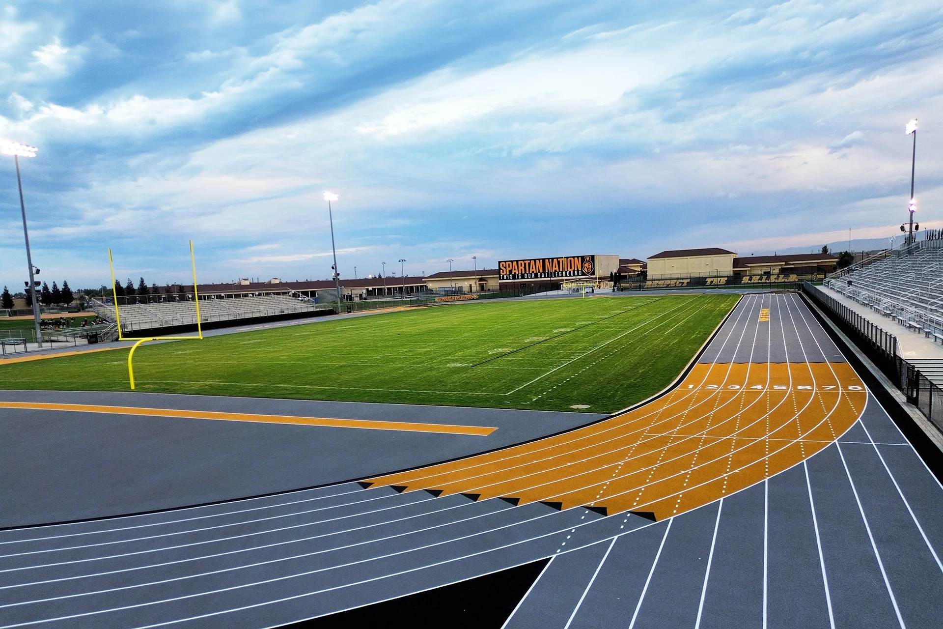 Ground-level view of a blue-grey rubberized running track curve and yellow exchange zones beneath stadium lighting and a cloudy sunset sky.