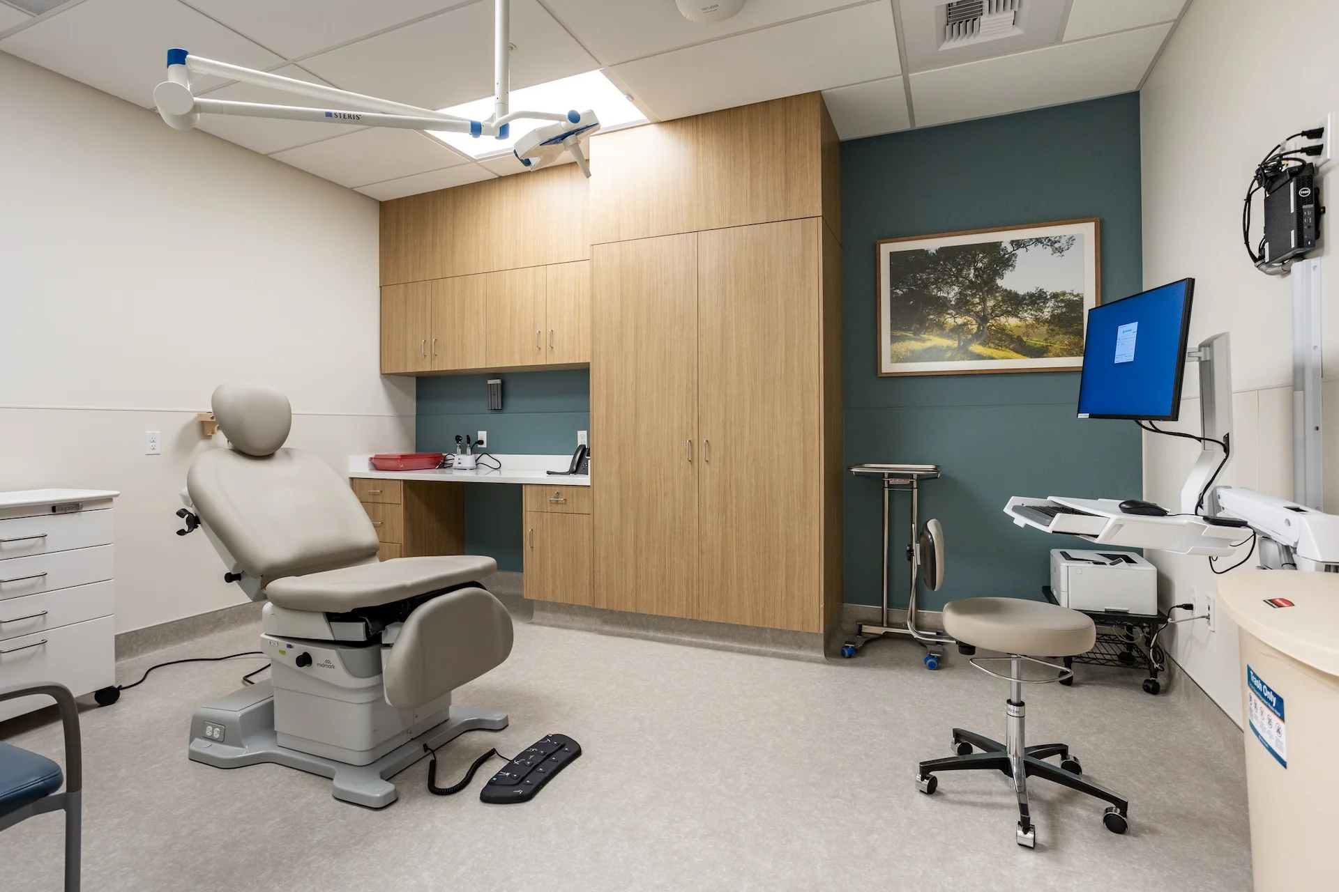 High-end clinical examination space featuring a motorized podiatry or dermatology procedure chair with foot controls, integrated wood veneer cabinetry, a ceiling-mounted adjustable examination lamp, a wall-mounted charting monitor, and a framed landscape picture.