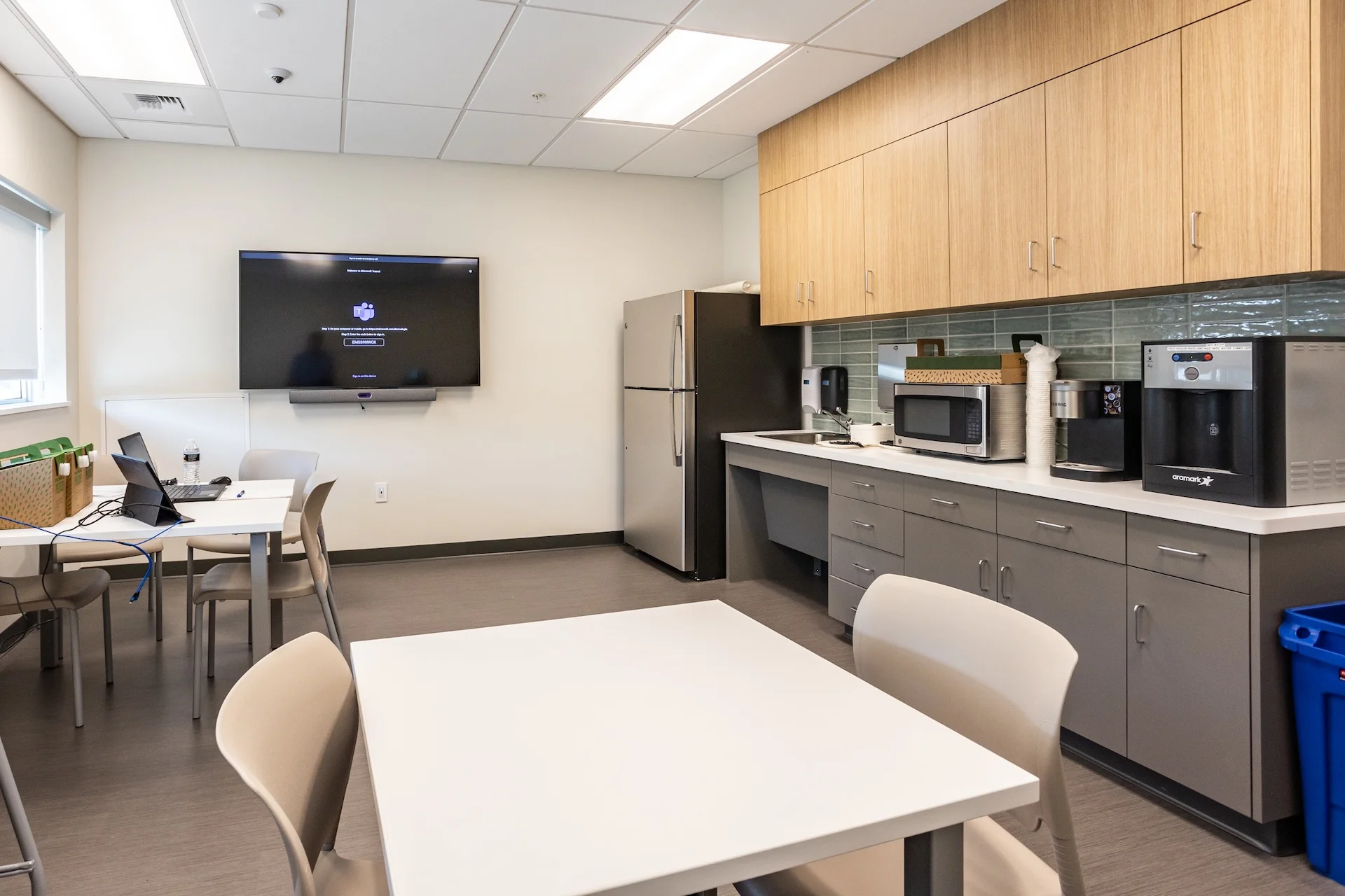 Modern institutional breakroom and dining space featuring upper wood grain cabinets and lower charcoal grey cabinetry, a custom glass tile backsplash, stainless steel refrigerator and microwave appliances, commercial coffee makers, an interactive smart monitor panel, and laminate white dining tables.
